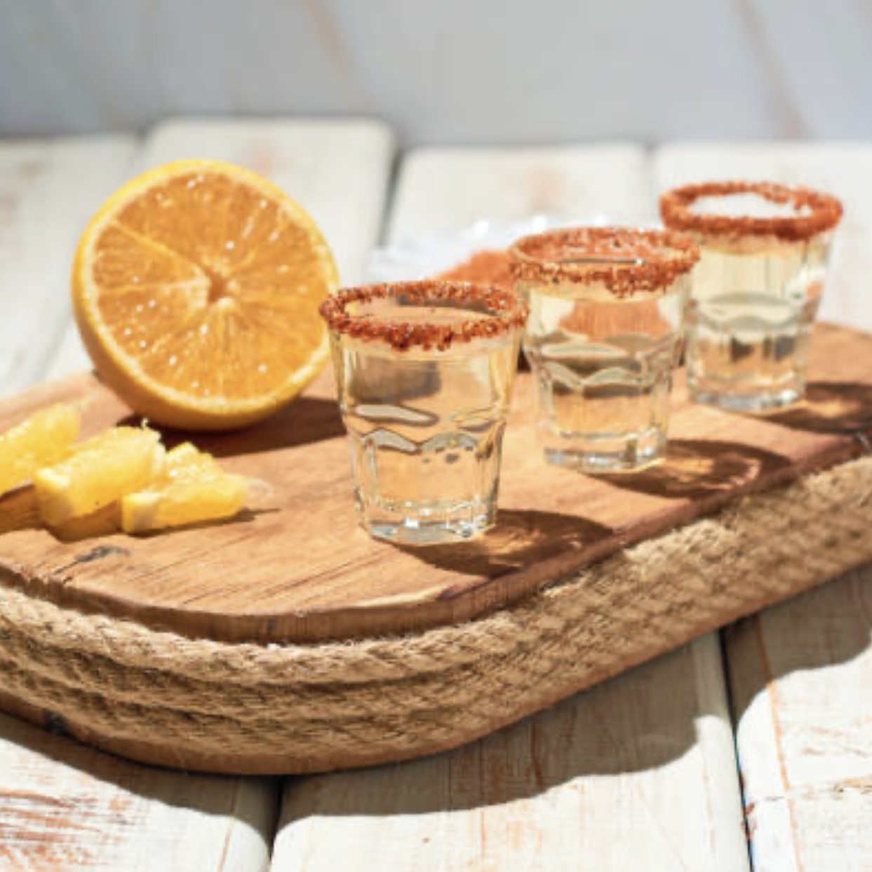 Three tequila shots with salted rims on a wooden tray, sliced lemon, and a halved lemon in the background sunlight on a white surface.