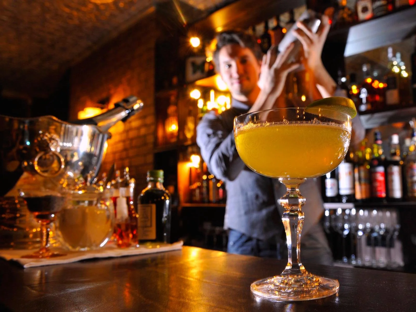 A yellow cocktail garnished with a lime wedge on a fancy glass in the foreground of a bar scene. A bartender is blurred in the background, holding a shaker. The bar has bottles of liquor and warm lighting.