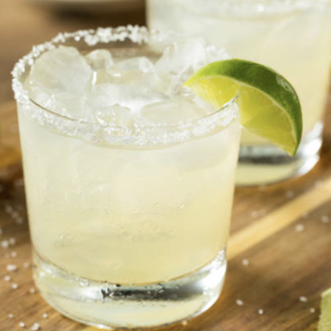 Close-up of a glass of margarita with a salted rim and lime wedge garnishment, filled with ice and pale yellow liquid, on a wooden surface.