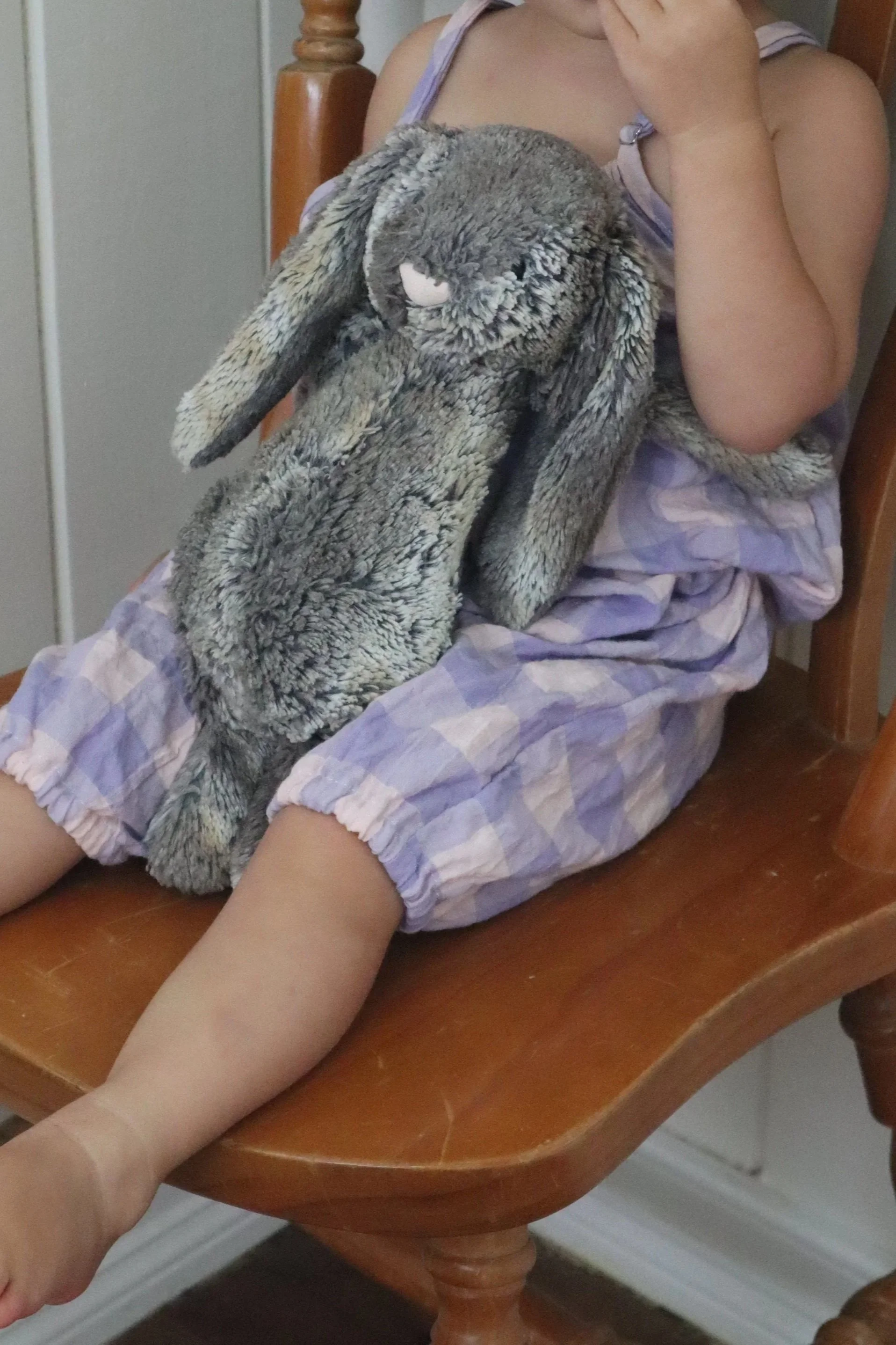 A young child sitting on a wooden chair wearing purple checkered pajamas, holding a large stuffed gray rabbit with long ears, while covering their face with one hand.