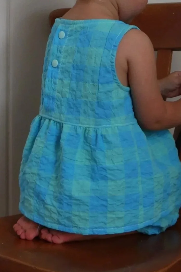 A young child sitting with knees folded on a wooden chair, wearing a light blue and green checked sleeveless dress, viewed from the back.