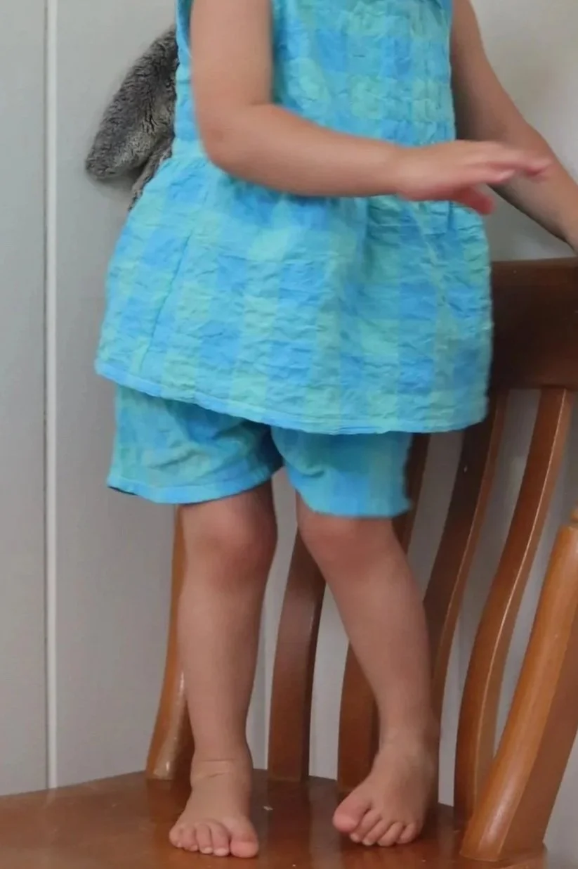 A child standing on a wooden chair, wearing a blue and green patterned sleeveless top and matching shorts, with a grey furry backpack behind them.