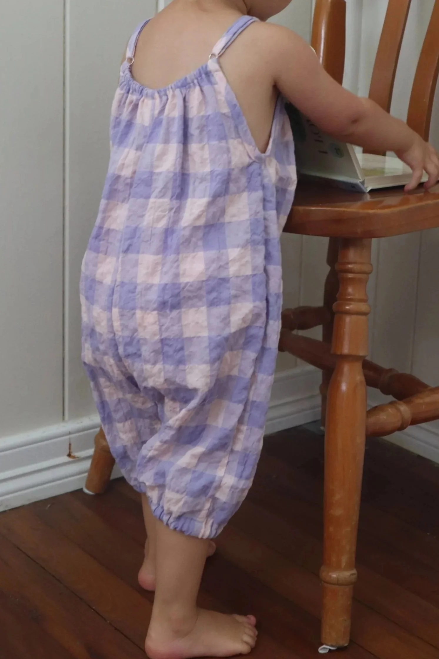 A young child standing barefoot next to a wooden table, wearing a loose, pastel-colored, checkered jumpsuit, reaching towards a book on the table.