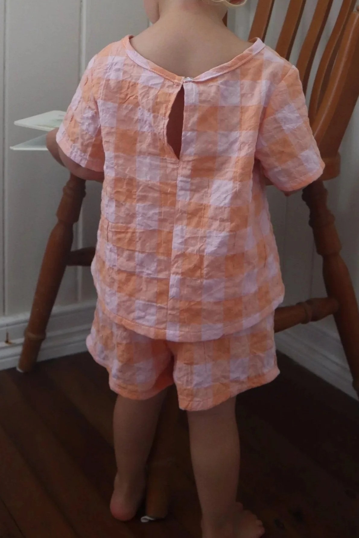 A child in pink and orange checkered pajamas standing in front of a wooden chair indoors.