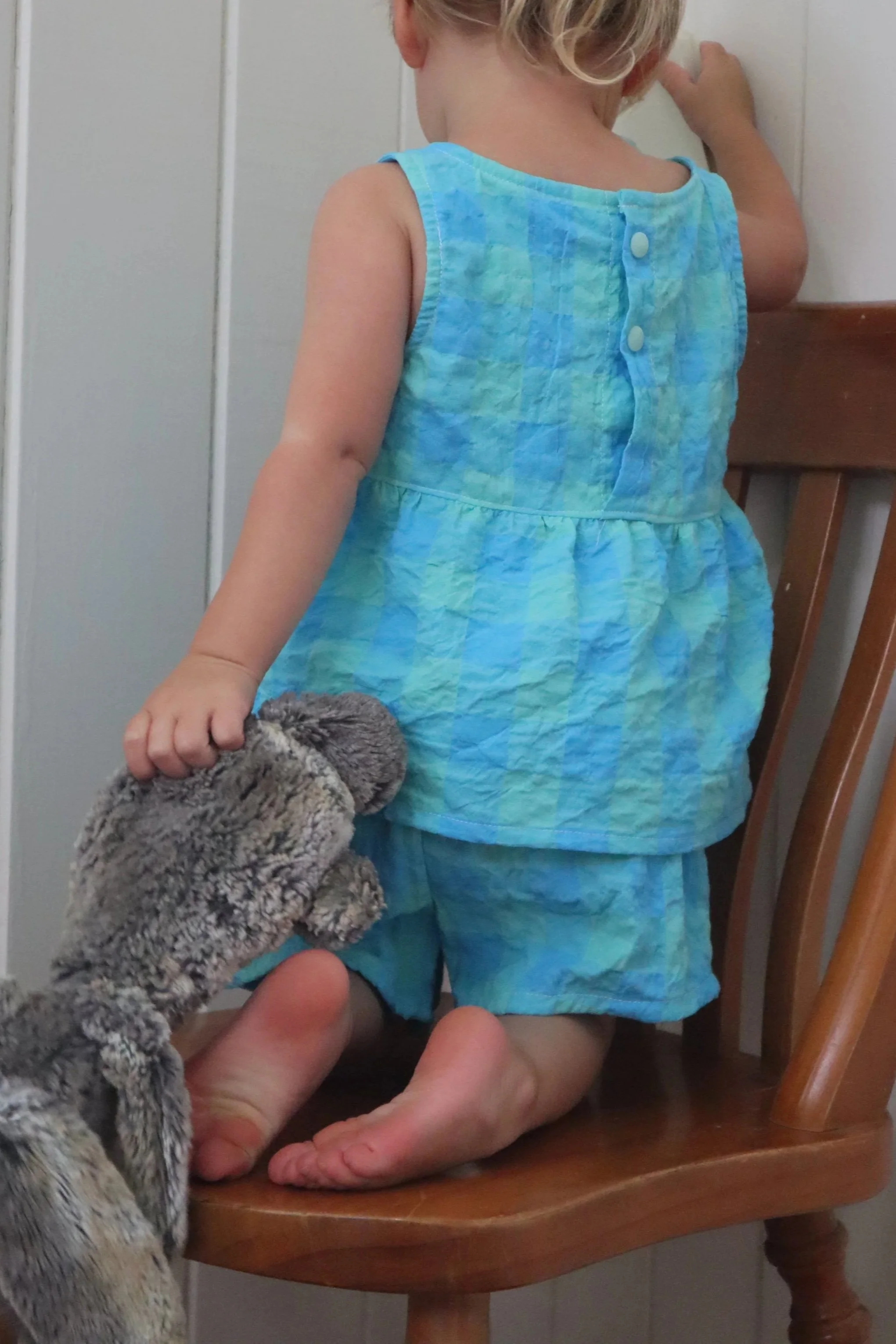 A young girl dressed in a blue-green plaid sleeveless top and matching shorts kneels on a wooden chair facing a wall, while a plush stuffed animal is held by a hand, with the girl's hand reaching toward the wall.