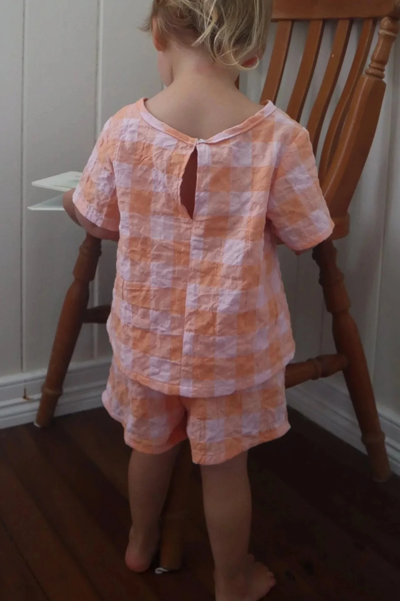 A young girl with blonde hair is wearing pink and white checkered pajamas, standing with her back to the camera in a room with wooden floors and furniture.