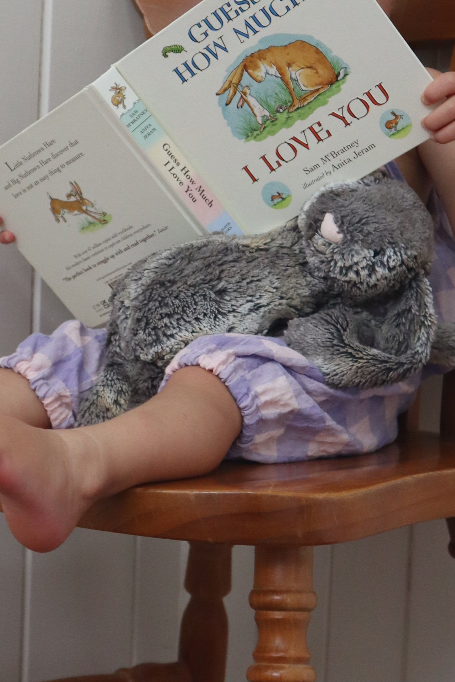 Child sitting on a wooden chair, holding a plush meerkat toy, with a children's book titled 'Guess How Much I Love You' in front of their face.