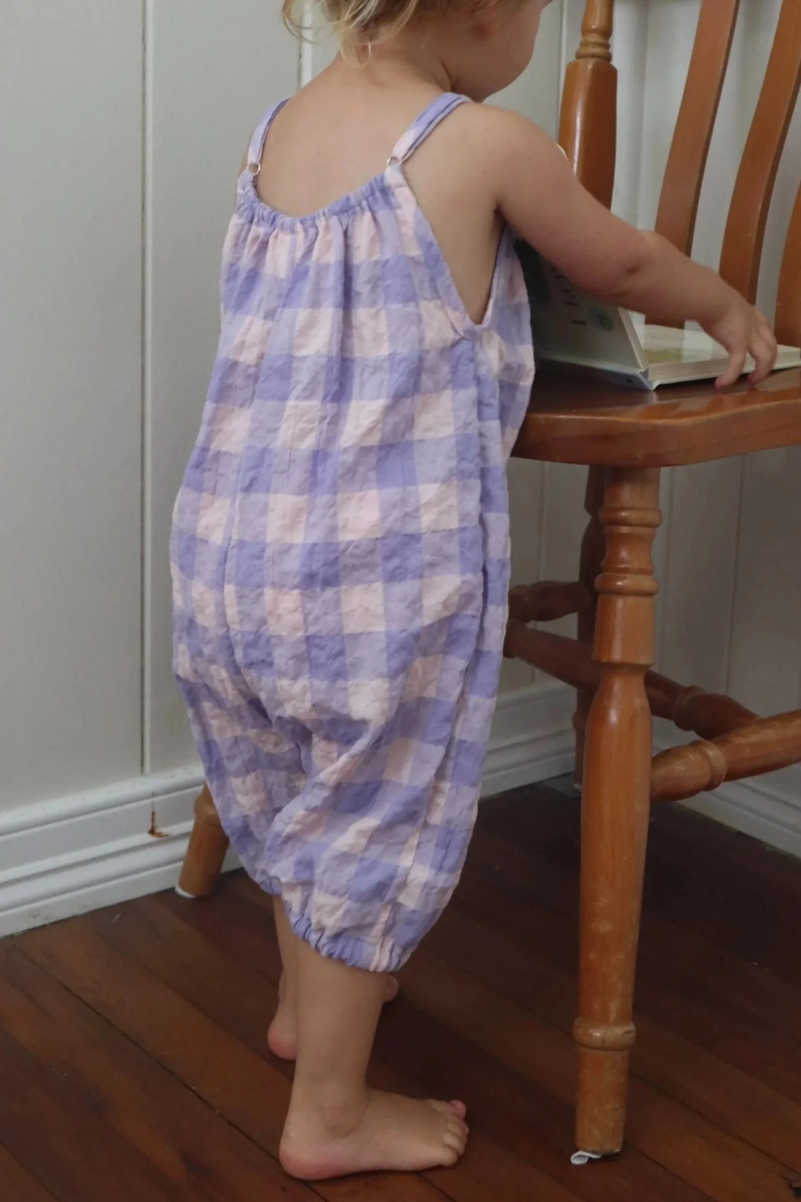 A young child standing barefoot next to a wooden chair, reaching for a book on the seat, wearing a purple and pink checkered sleeveless jumpsuit.