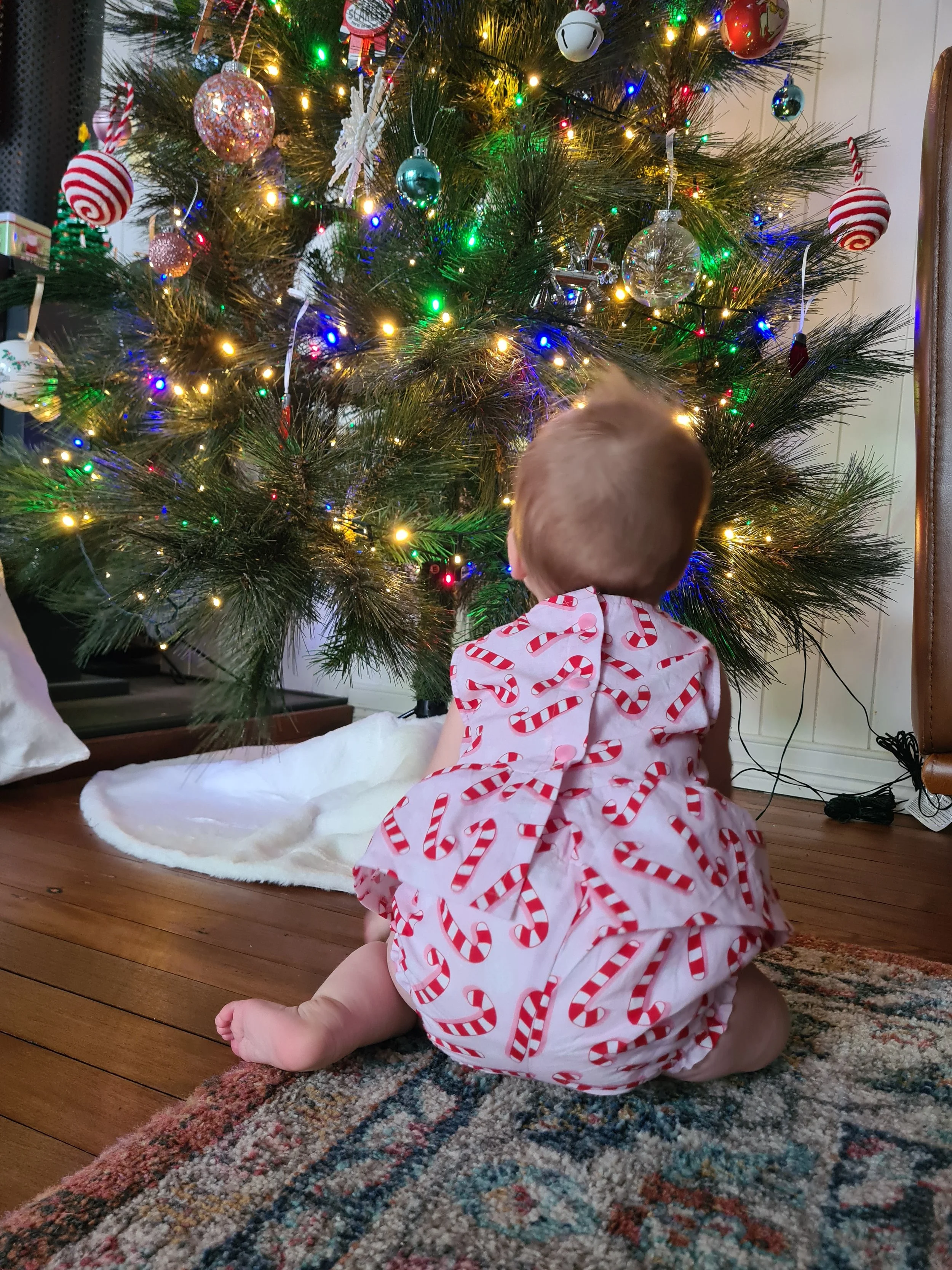 A young child sitting on a rug facing a decorated Christmas tree with colorful lights and ornaments, wearing a white outfit with red candy cane patterns.
