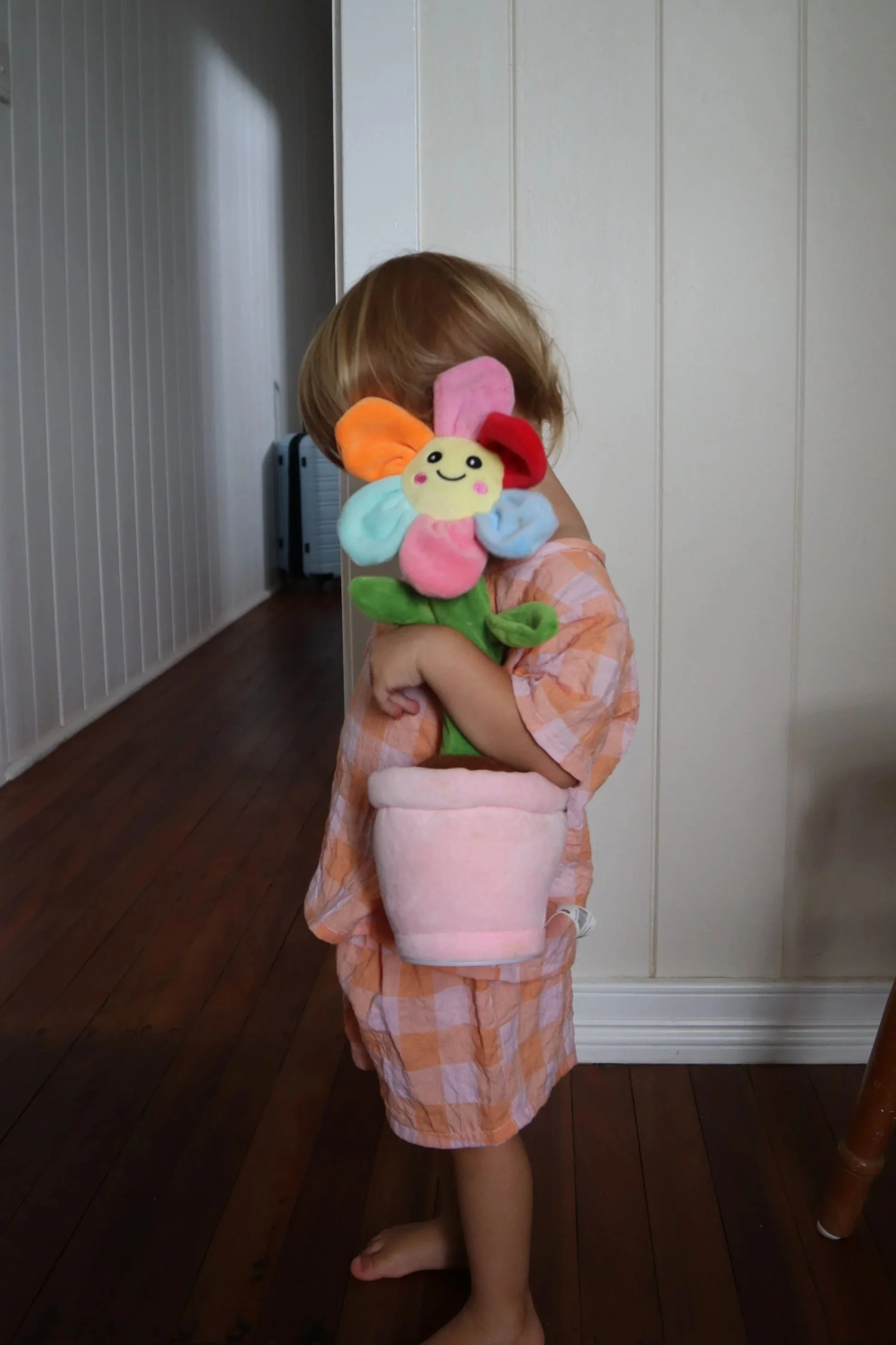 A young child standing barefoot on a wooden floor, holding a plush flower toy with a smiling face, in front of a white wall in a home interior.