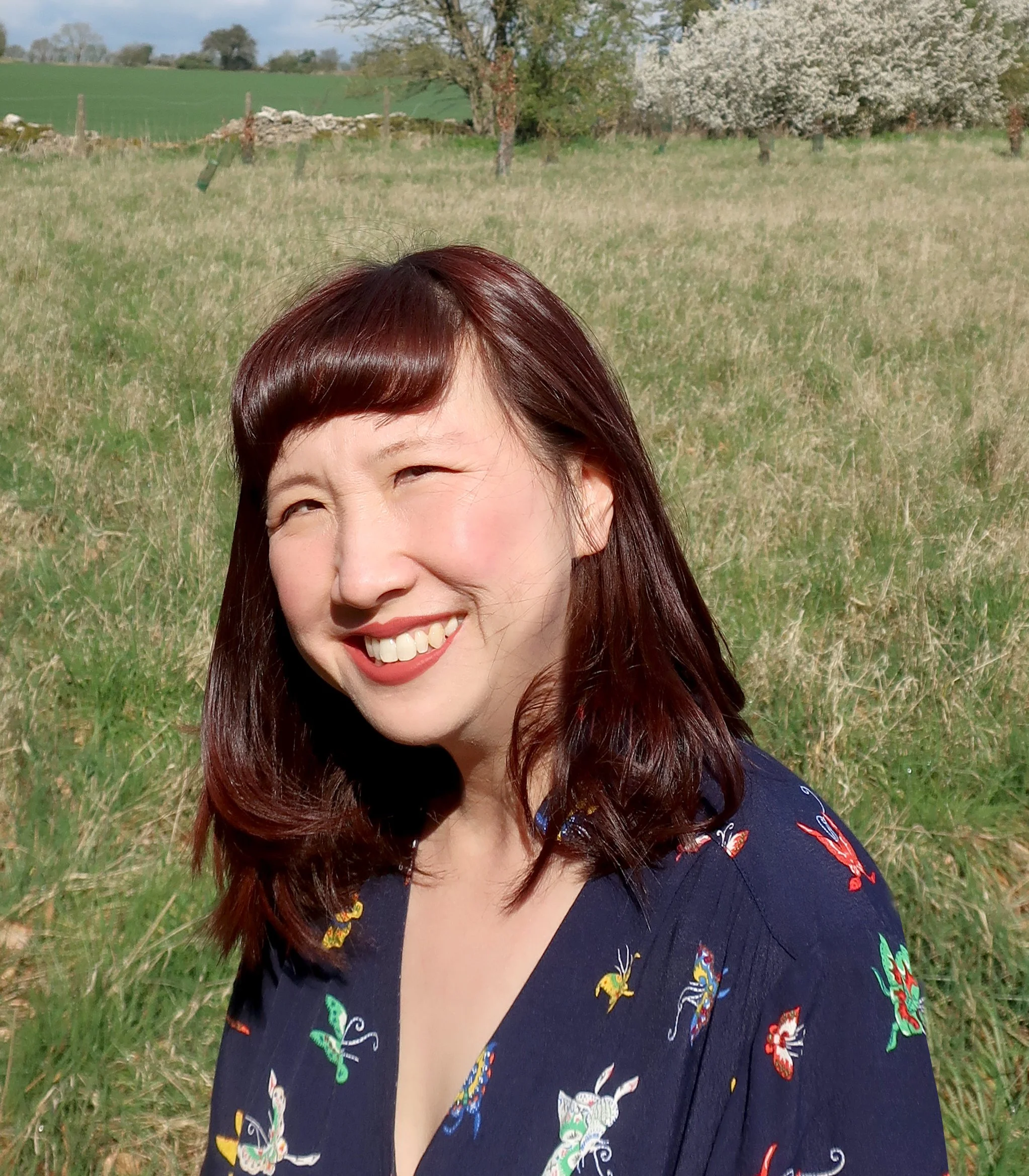 A woman with shoulder-length red hair, smiling, outdoors in a grassy field with trees and a stone wall in the background.