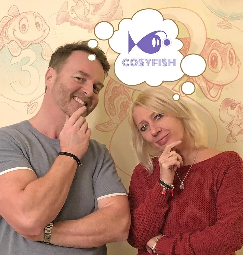 A man and woman posing with thinking poses in front of a background with cartoon fish drawings, including a frog and other aquatic creatures. The man is smiling and wearing a gray t-shirt, the woman is wearing a red sweater. A thought bubble above her head contains a logo with a purple fish and the word "COSYFISH."