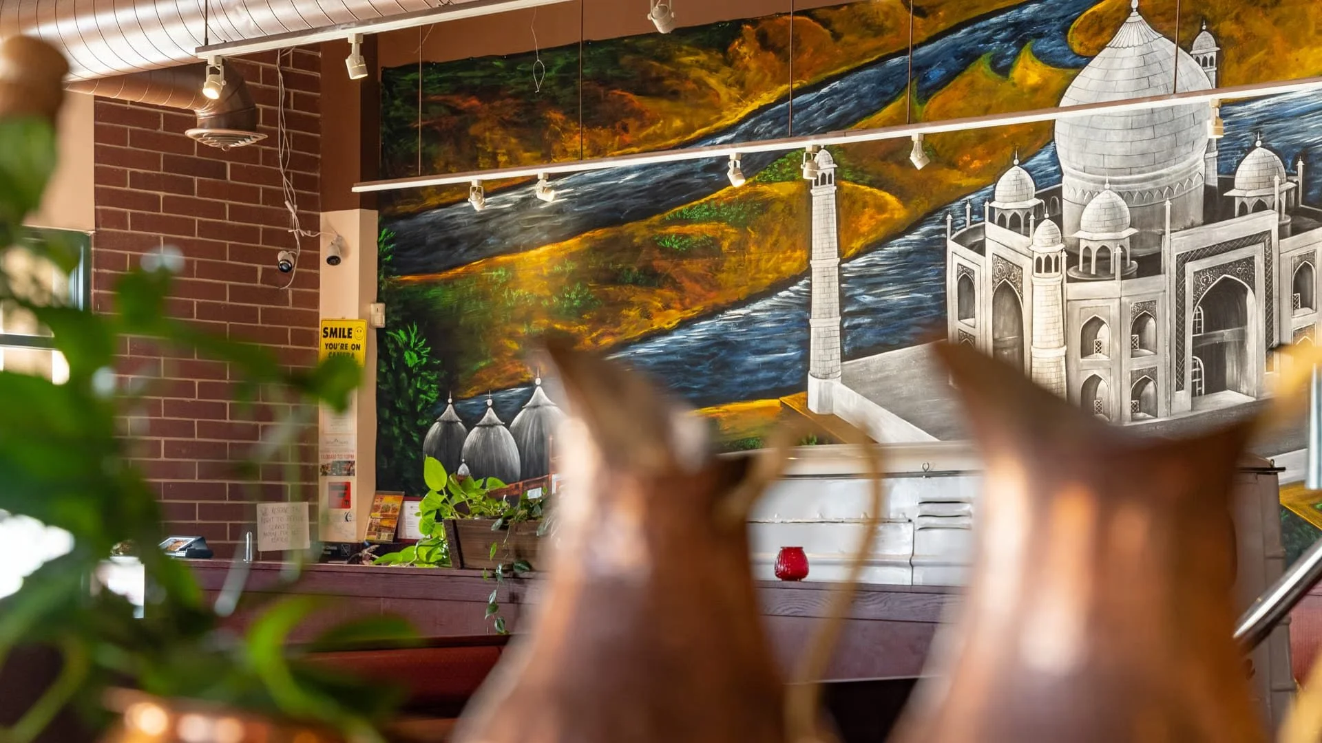 Interior of a restaurant or cafe with a large mural of the Taj Mahal in the background, surrounded by greenery and a flowing river, with a brick wall on the left, potted plants, and blurred objects in the foreground.