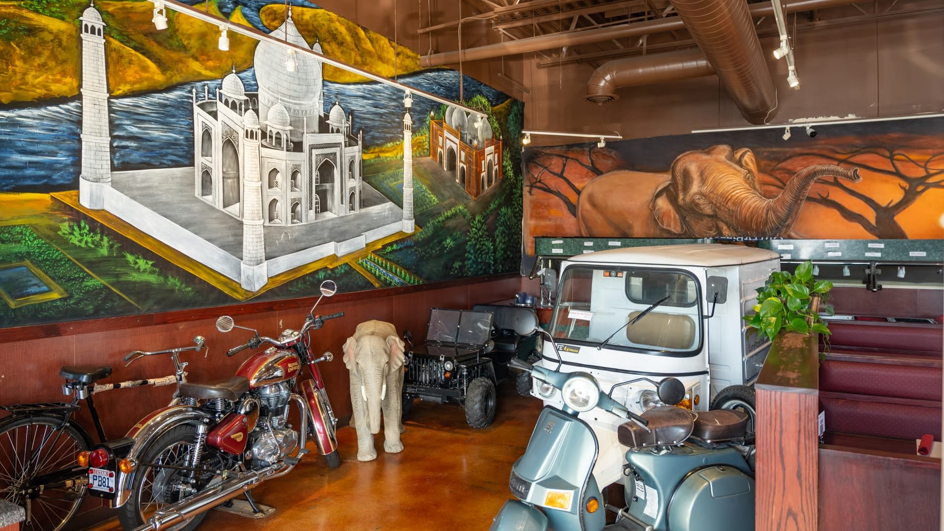 Interior of a museum with colorful murals, including the Taj Mahal, a landscape with an elephant, and a safari scene. Several vintage vehicles are displayed, including a red motorcycle, a small black off-road vehicle, a white scooter, and a blue scoo