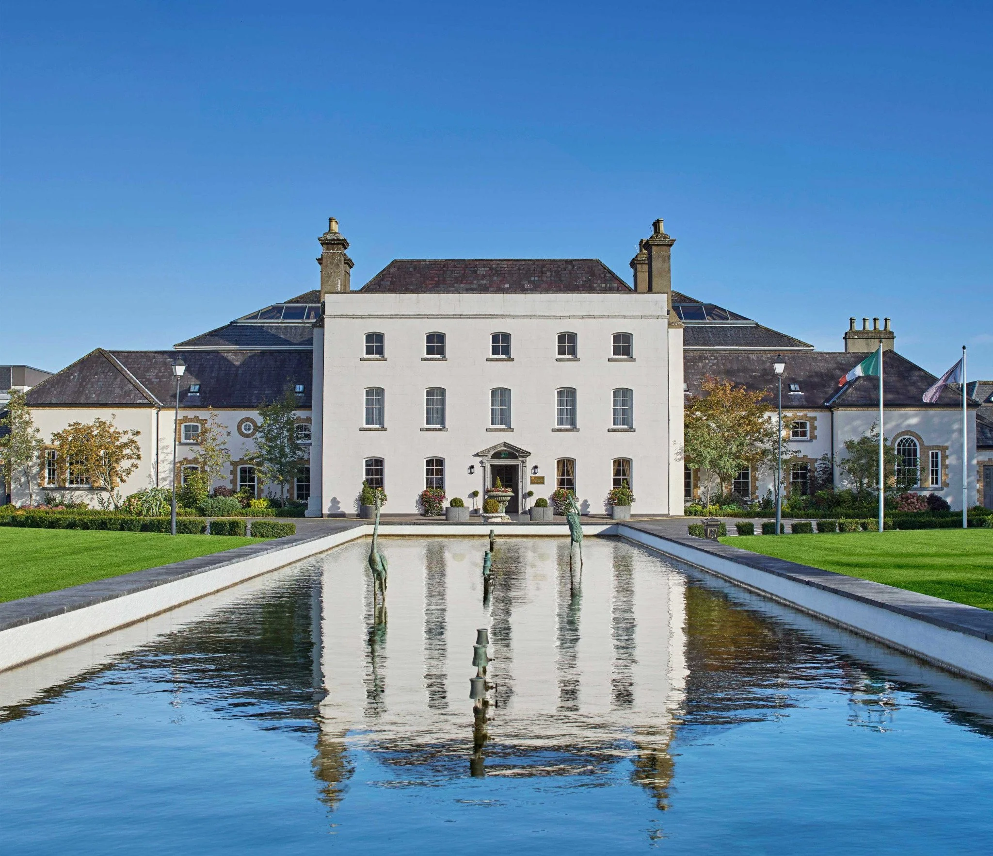 A white multi-story building with a sloped roof and chimney stacks, complemented by a reflecting pool with sculptures in the foreground, surrounded by green trees and a clear blue sky.