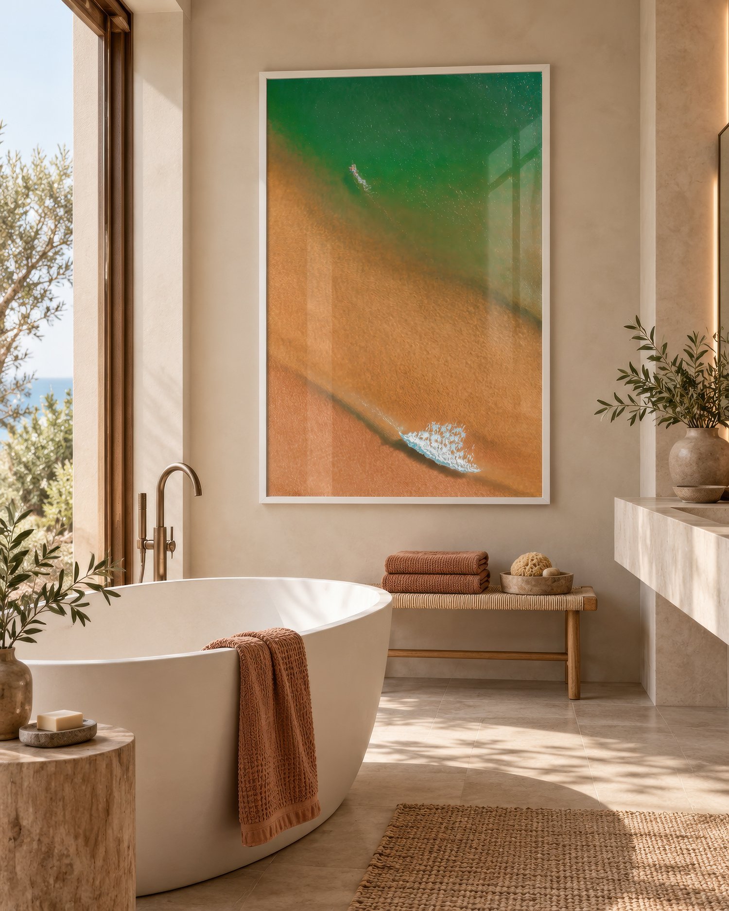 Aerial coastal photograph of green ocean water meeting golden sand, displayed in a white frame above a freestanding bathtub in a sunlit bathroom with natural stone and timber accents, by Nick Psomiadis at NIKART Studio and Gallery