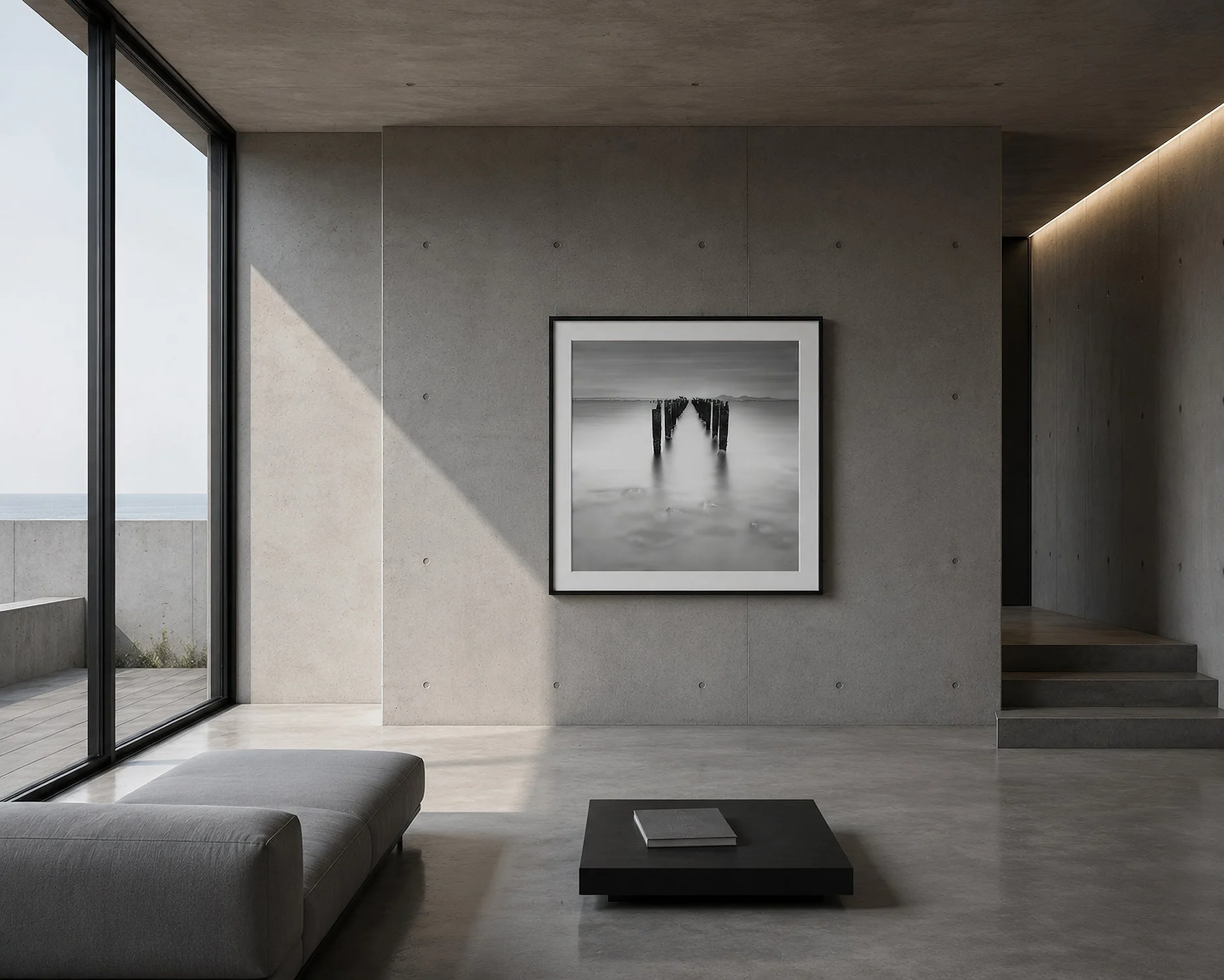 Minimalist concrete interior with a framed black and white jetty photograph on a raw concrete wall, styled with a low grey sofa, black coffee table, polished concrete floor and strong natural light, by Nick Psomiadis at NIKART Studio and Gallery.