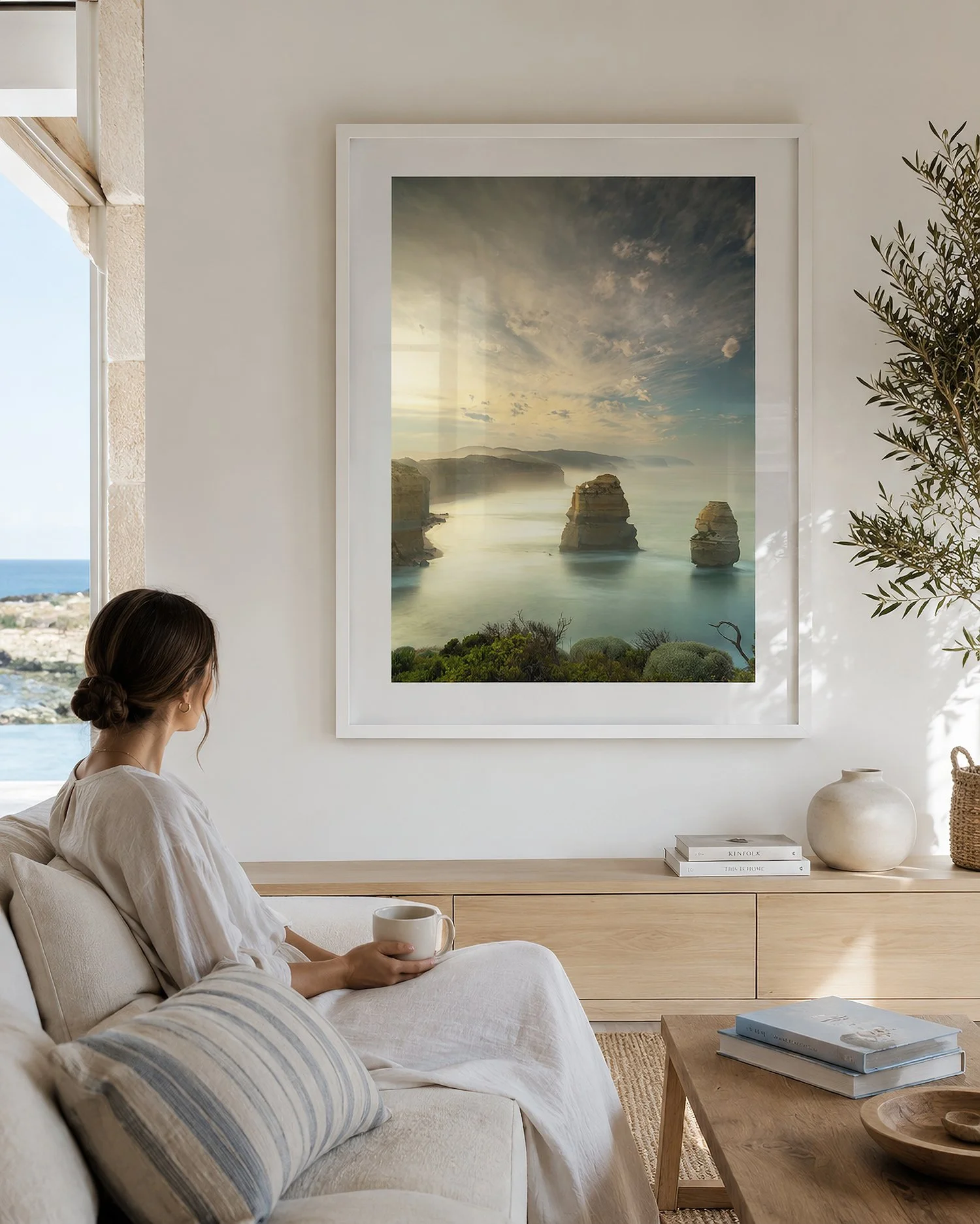Bright coastal living room interior with a seated woman beside a white-framed photograph of the Gog and Magog limestone stacks at Gibson Steps, with soft reflections on the glass, by Nick Psomiadis at NIKART Studio and Gallery.