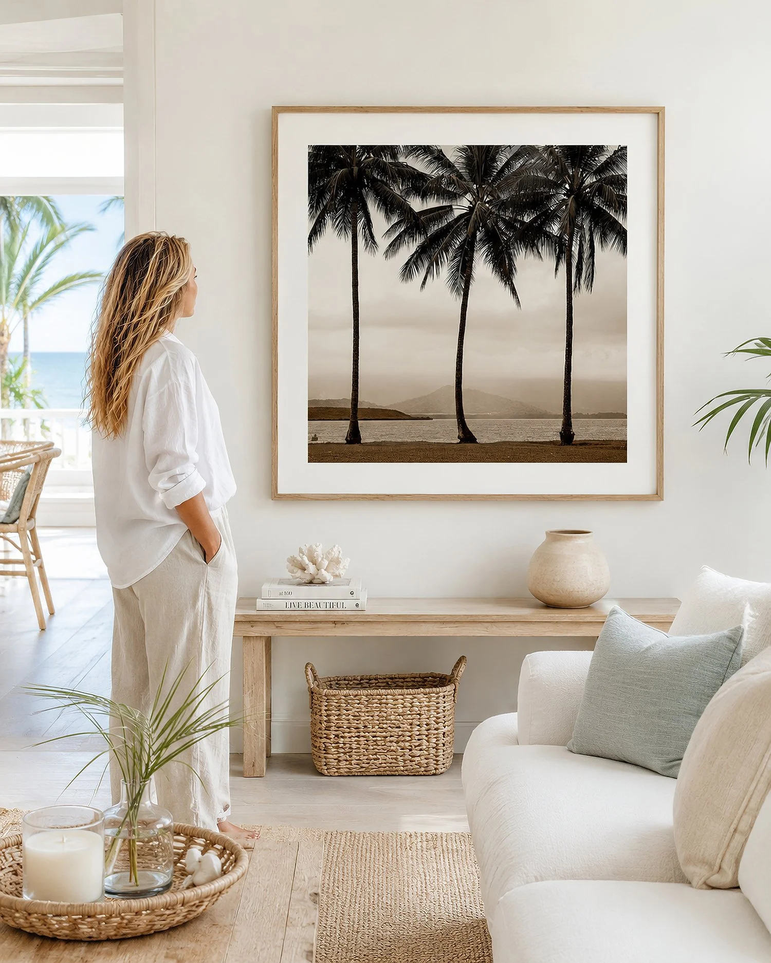 Interior photograph of a bright coastal living room with framed palm tree artwork and seated woman.