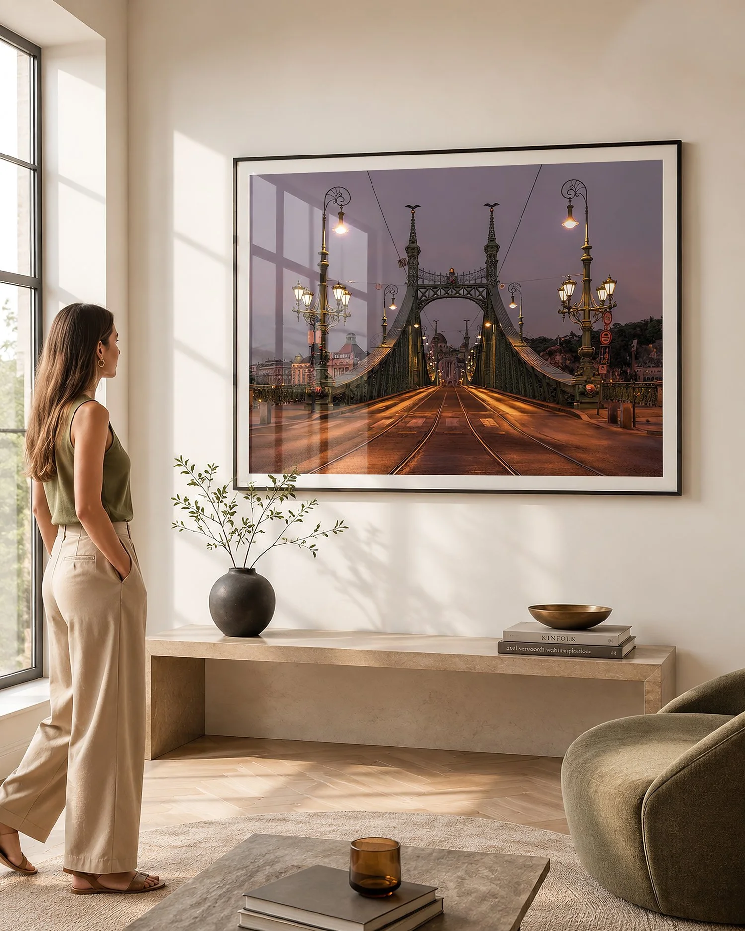 Bright minimalist interior with a woman viewing a framed photograph of Budapest’s illuminated Liberty Bridge, styled with warm stone, olive seating and subtle glass reflections by Nick Psomiadis at NIKART Studio and Gallery