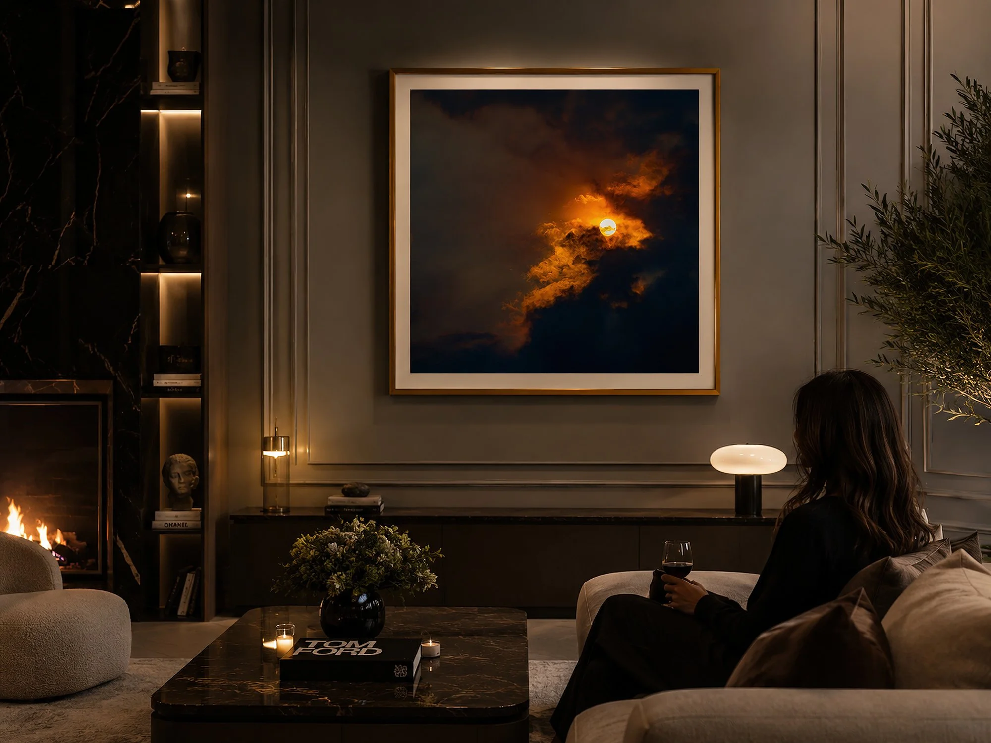 Seated woman in a luxurious living room with a gold-framed sky photograph above a dark console, surrounded by warm lighting, marble accents and elegant furnishings by Nick Psomiadis at NIKART Studio and Gallery