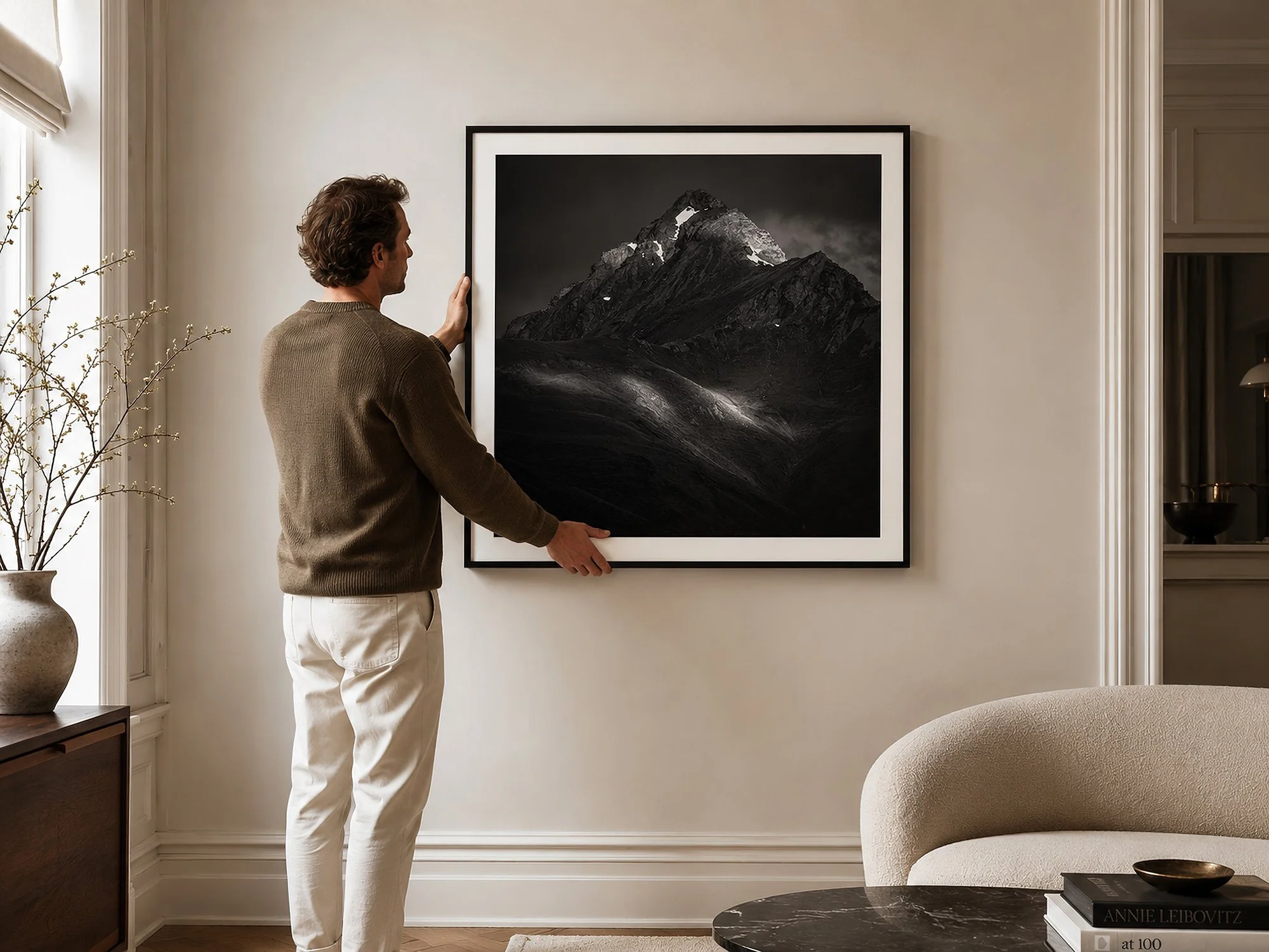 Man holding a square black-framed mountain photograph with a white border, preparing to hang it in a bright elegant living room by Nick Psomiadis at NIKART Studio and Gallery