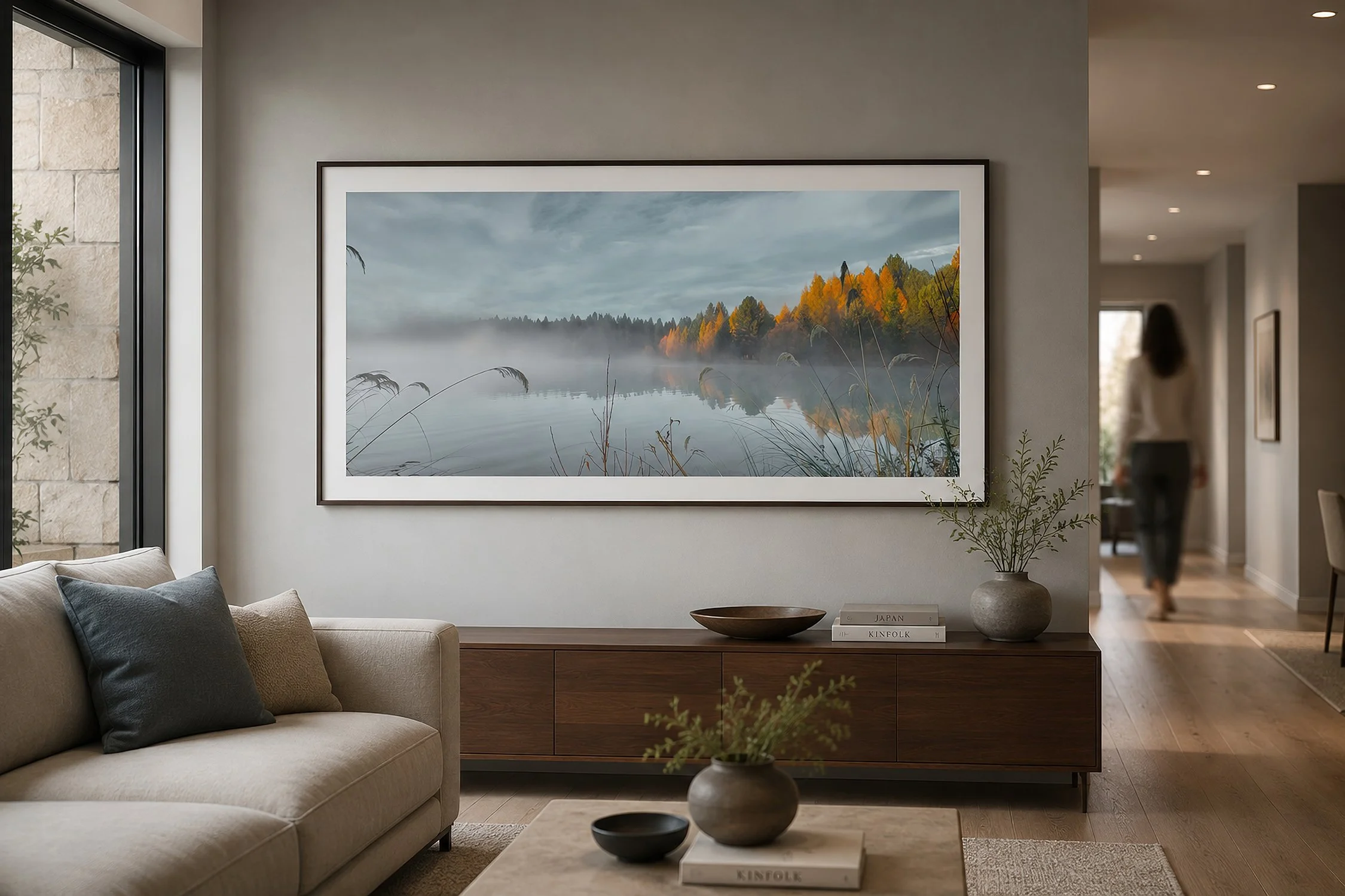 Modern living room with a framed misty autumn lake photograph and a person walking in the background, by Nick Psomiadis at NIKART Studio and Gallery.