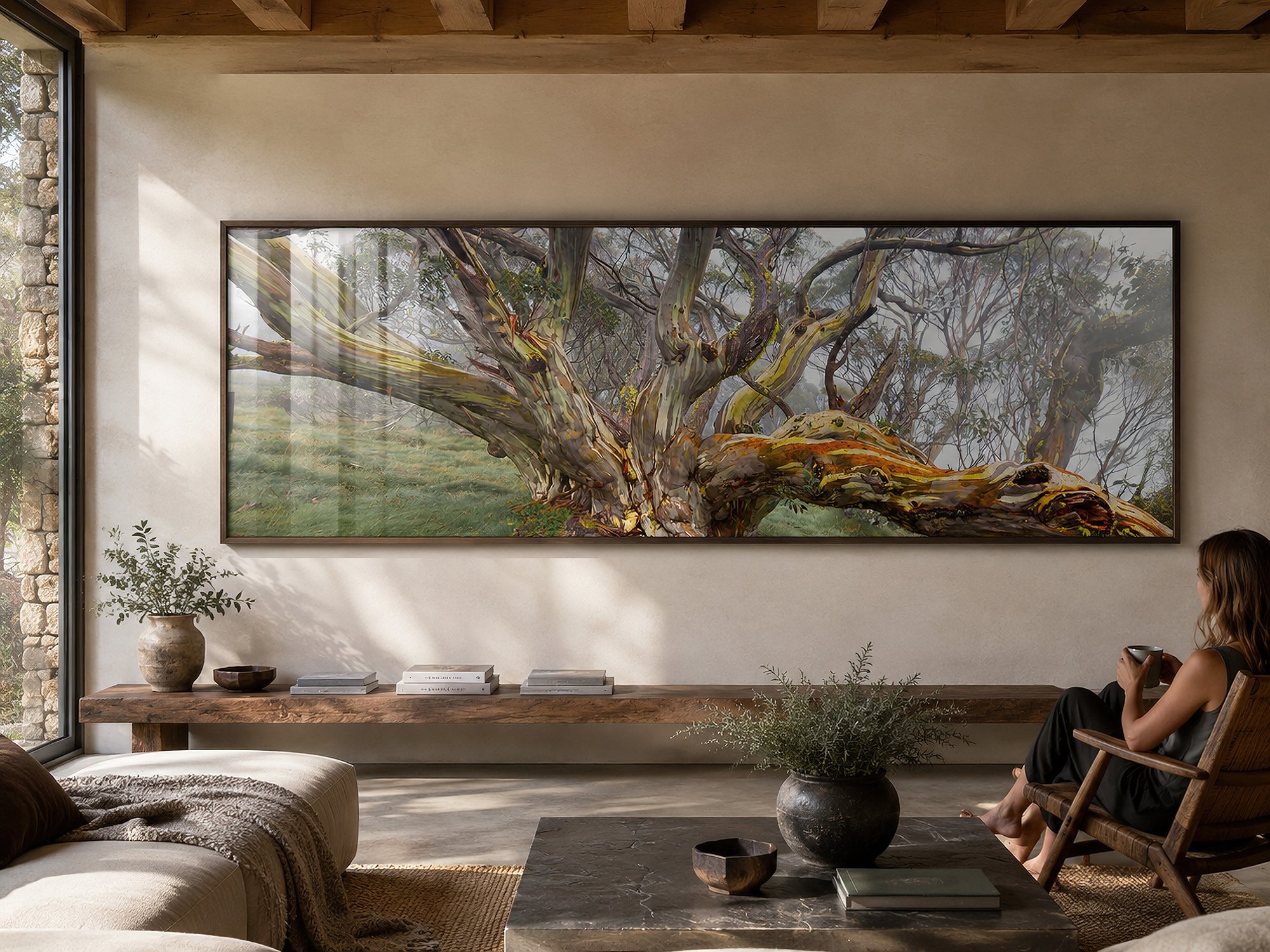 Bright moody living room with a seated woman beside a long black-framed panoramic snow gum landscape photograph with glass reflections above a timber bench by Nick Psomiadis at NIKART Studio and Gallery