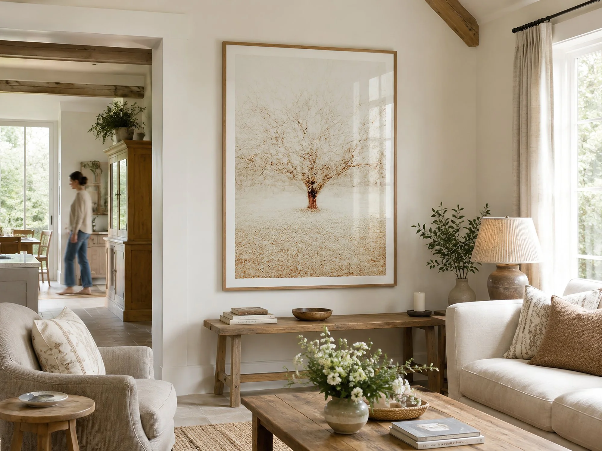 Bright country living room with a large framed sepia tree photograph, cream furnishings, rustic timber furniture, soft natural daylight and a person walking in the background, by Nick Psomiadis at NIKART Studio and Gallery