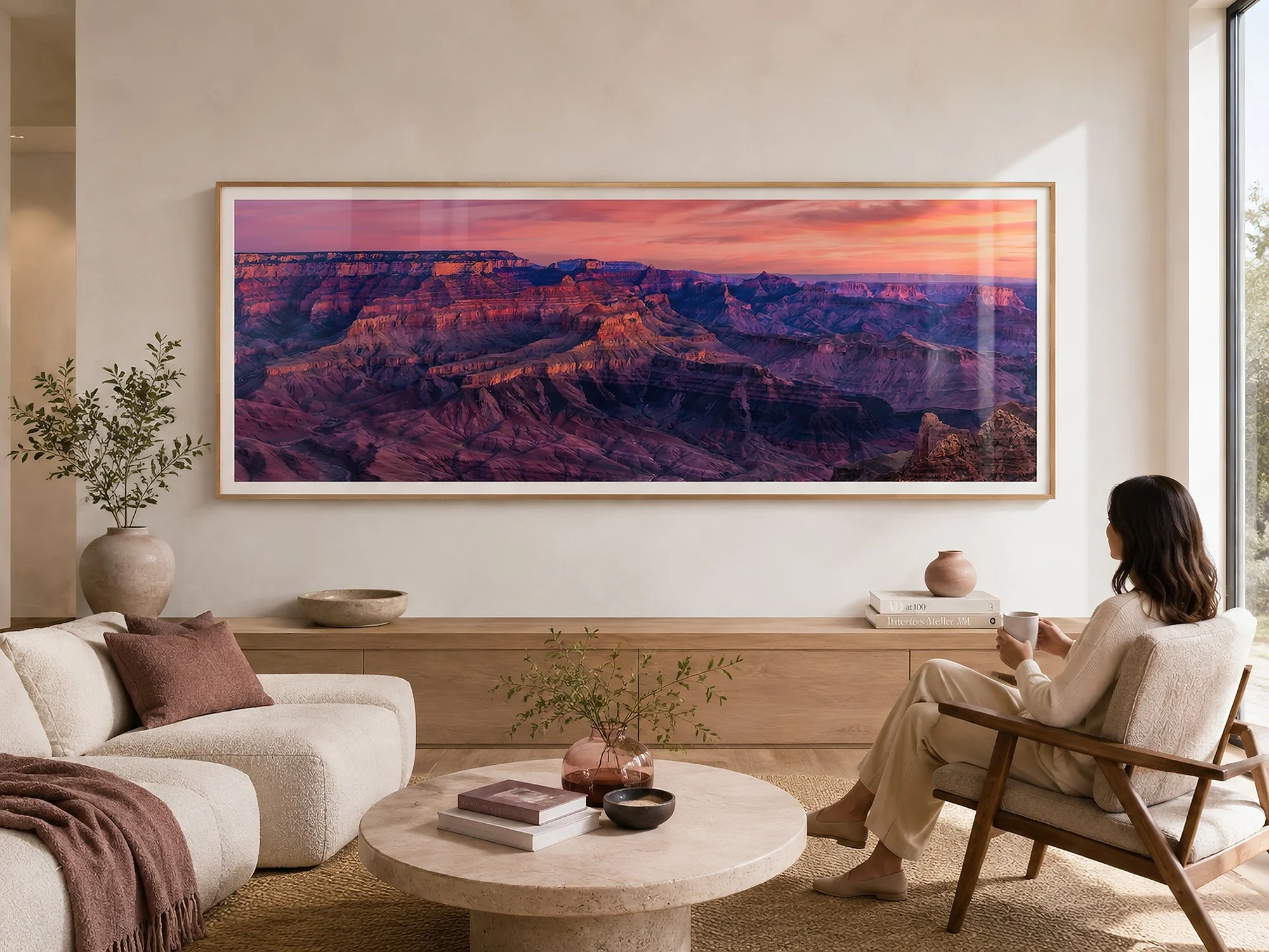 Framed panoramic Grand Canyon photograph with layered purple cliffs and a glowing pink sunset in a bright minimalist living room with neutral furnishings, soft rose accents and a seated woman, by Nick Psomiadis at NIKART Studio and Gallery