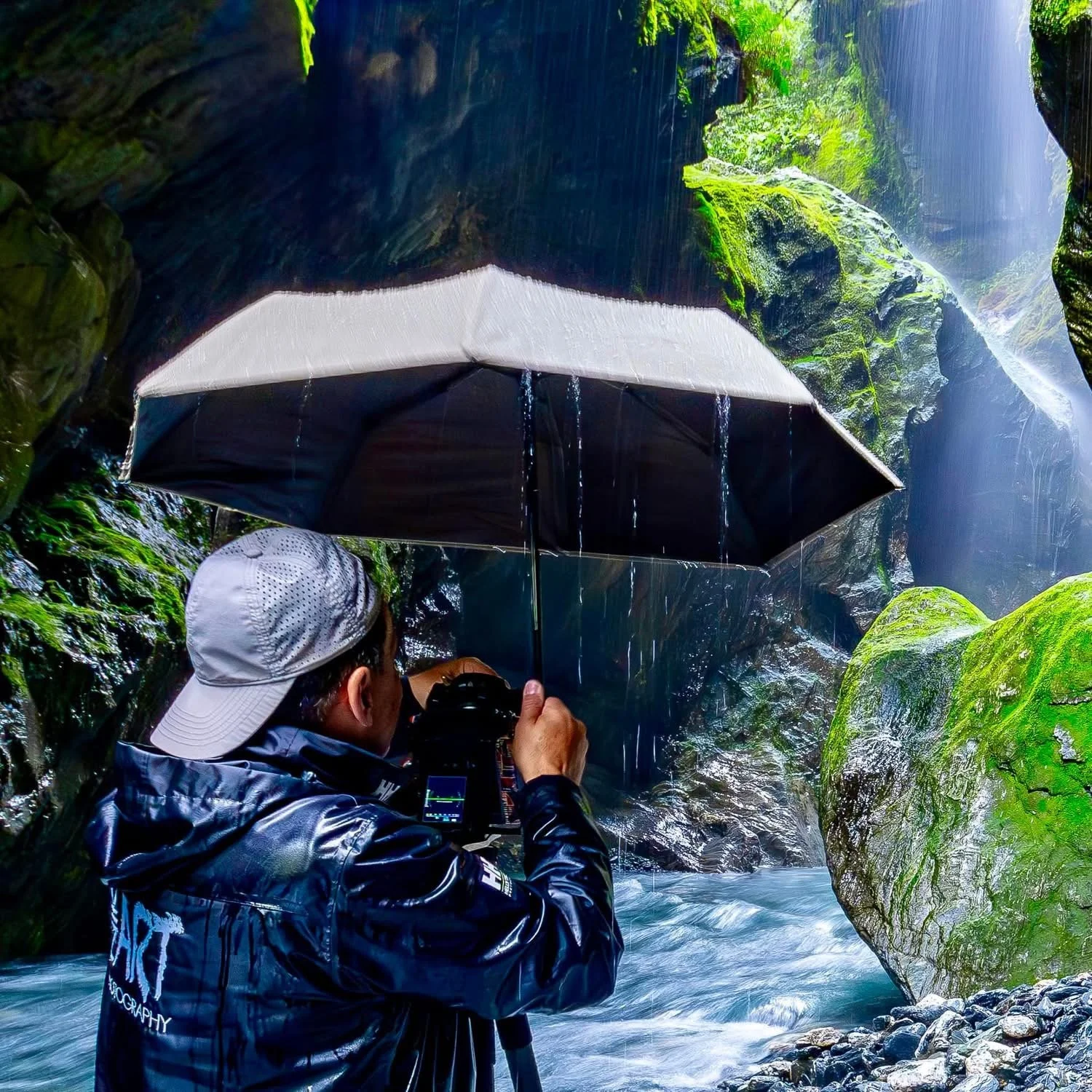 Photographer standing beneath umbrella in a moss canyon photographing waterfall landscape