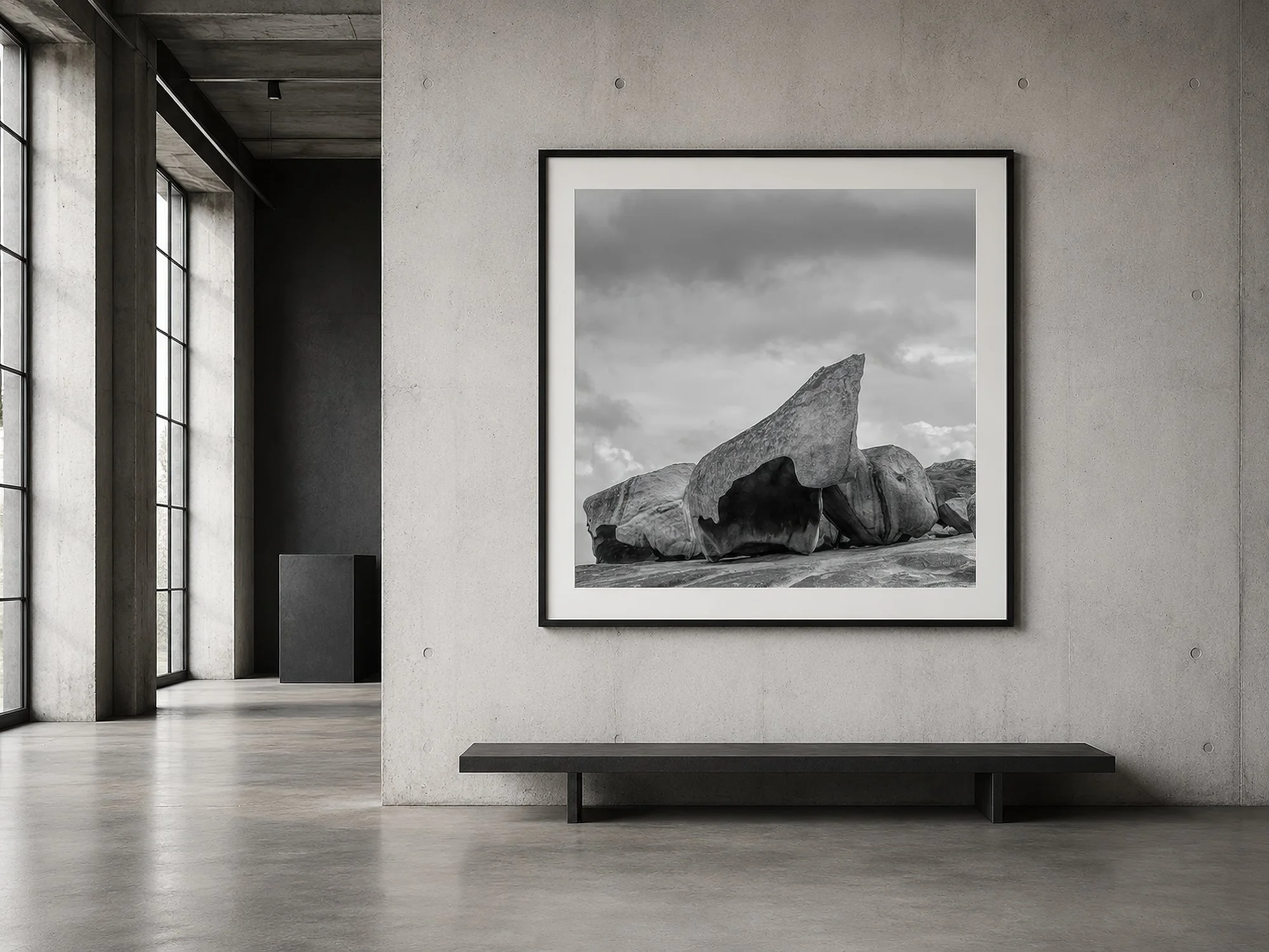 Framed black-and-white rock formation photograph in a sparse concrete warehouse with tall industrial windows and a minimalist bench by Nick Psomiadis at NIKART Studio and Gallery