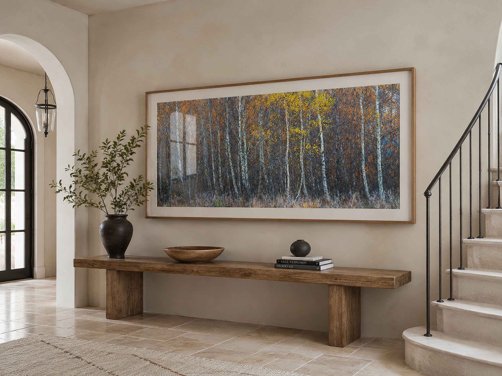 Large framed panoramic autumn forest photograph displayed in a bright neutral entry hall with a rustic timber console, curved staircase and arched doorway by Nick Psomiadis at NIKART Studio and Gallery