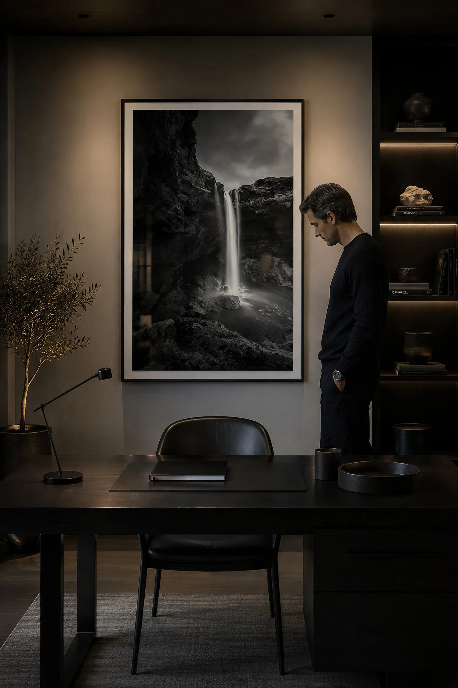 Moody modern minimalist study with a framed black-and-white photograph of Kvernufoss waterfall, dark timber desk, warm shelf lighting and a man standing beside the desk looking down, by Nick Psomiadis at NIKART Studio and Gallery