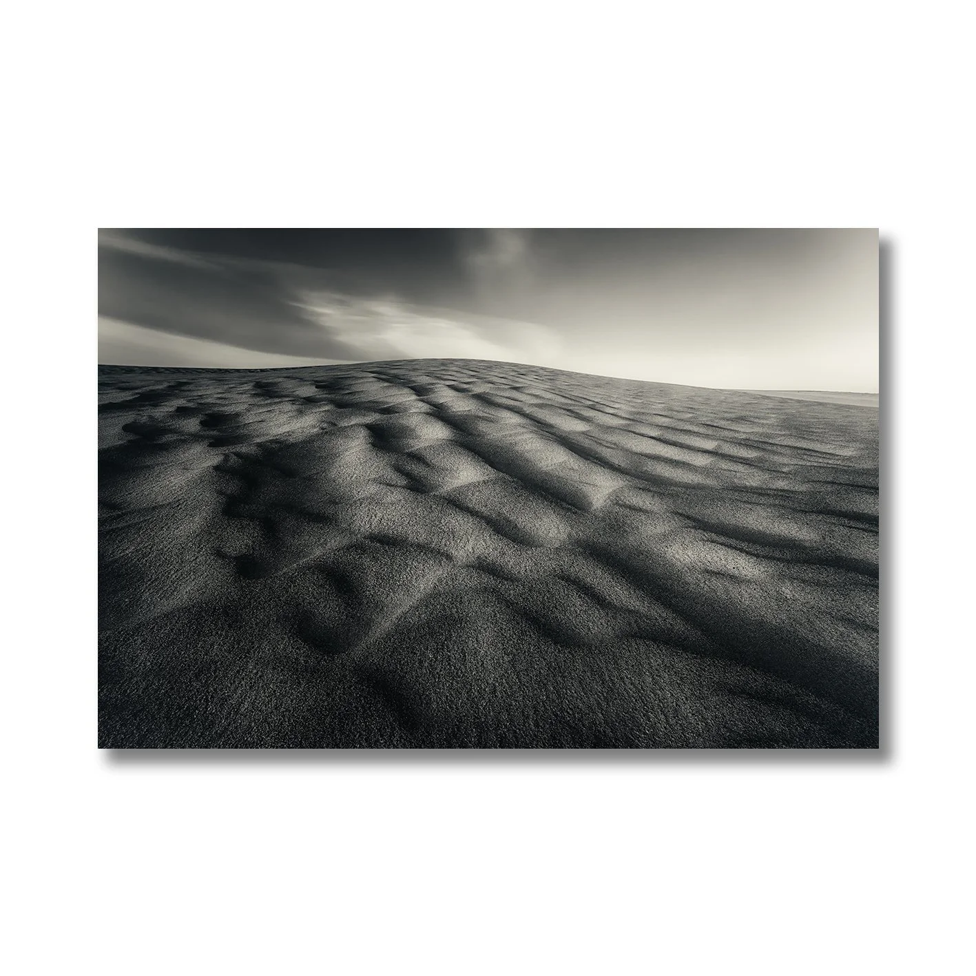 Black and white photograph of wind-sculpted sand dunes beneath a dramatic sky.