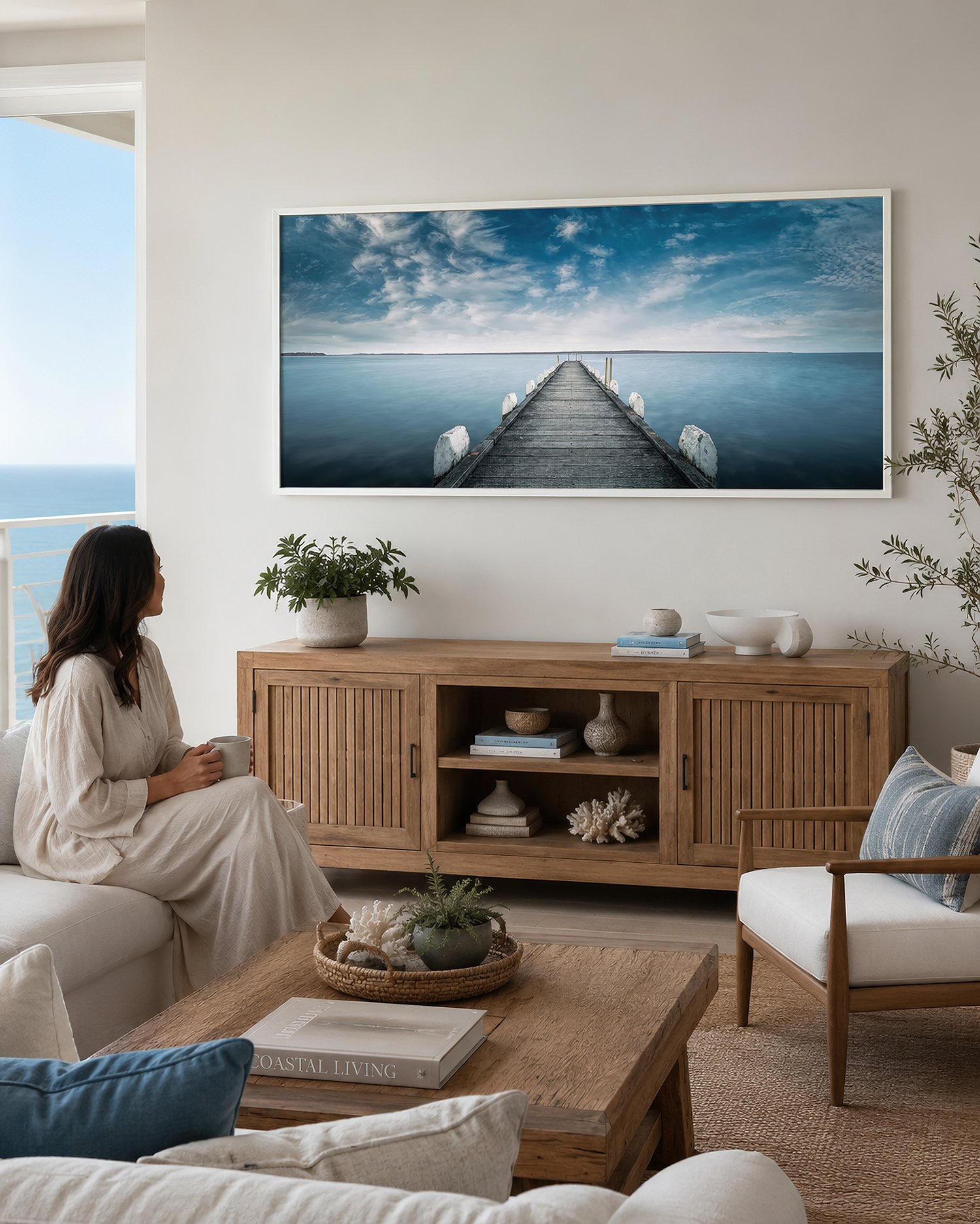 Coastal lounge featuring a white-framed canvas photograph of Grantville Jetty extending into calm blue water, with a seated woman admiring the artwork, by Nick Psomiadis at NIKART Studio and Gallery.