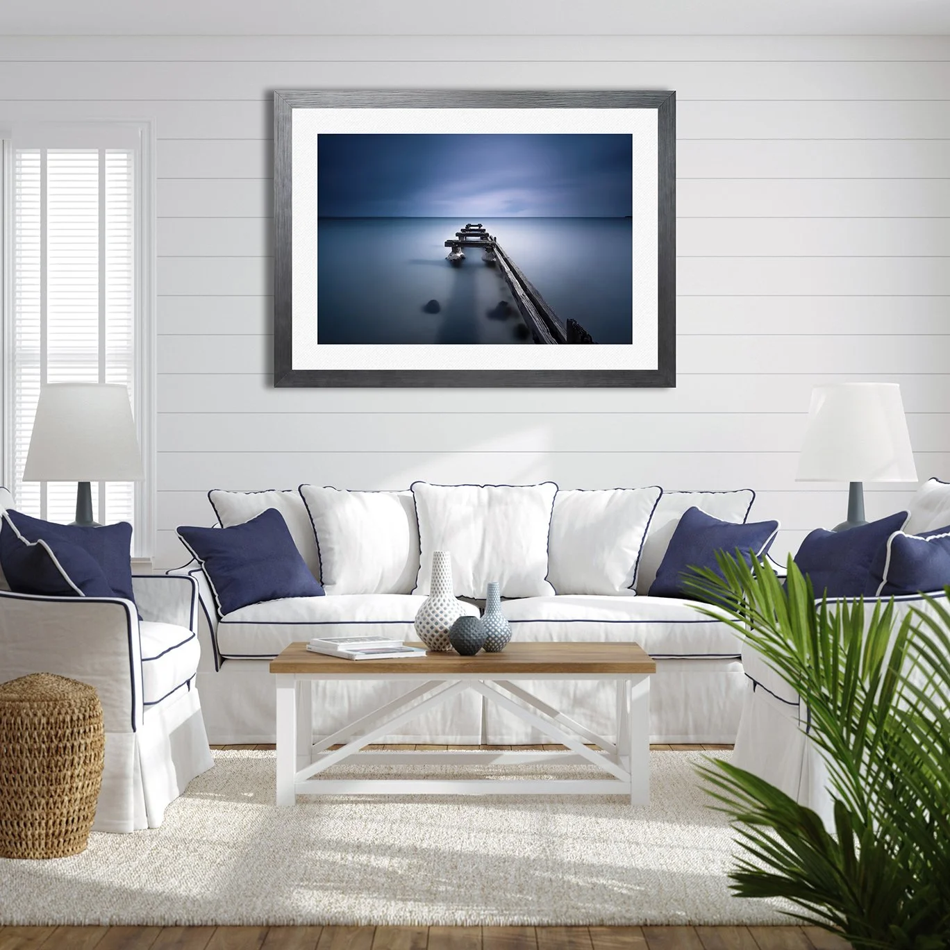 Living room with white walls, white sofa with navy pillows, a wooden coffee table, and a large framed photograph of a wooden pier leading into the ocean hanging on the wall.