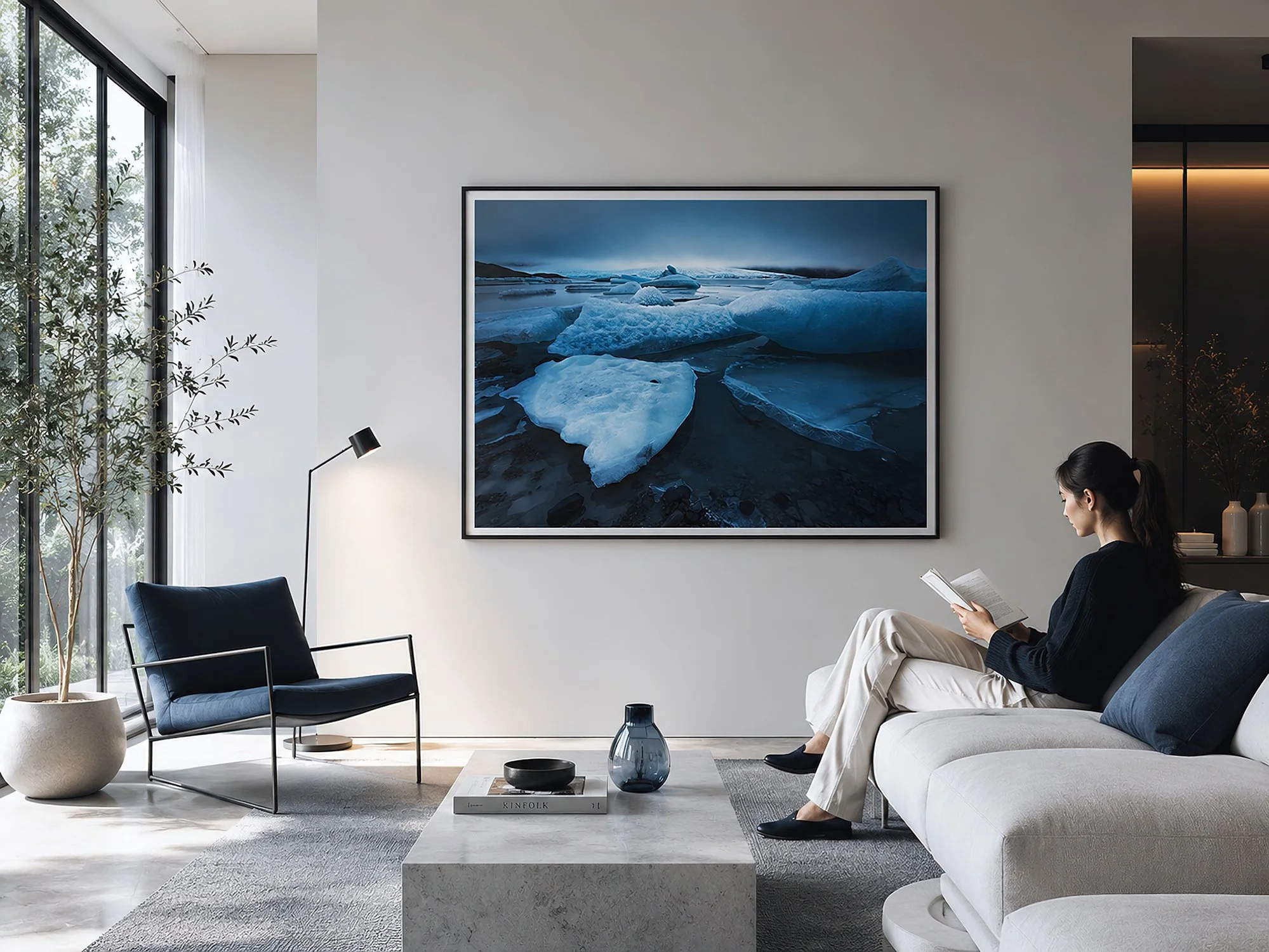 Bright modern minimalist living room with a framed blue glacial landscape photograph above a cream sofa, styled with navy accents, natural daylight, subtle warm lighting and a woman seated reading, by Nick Psomiadis at NIKART Studio and Gallery