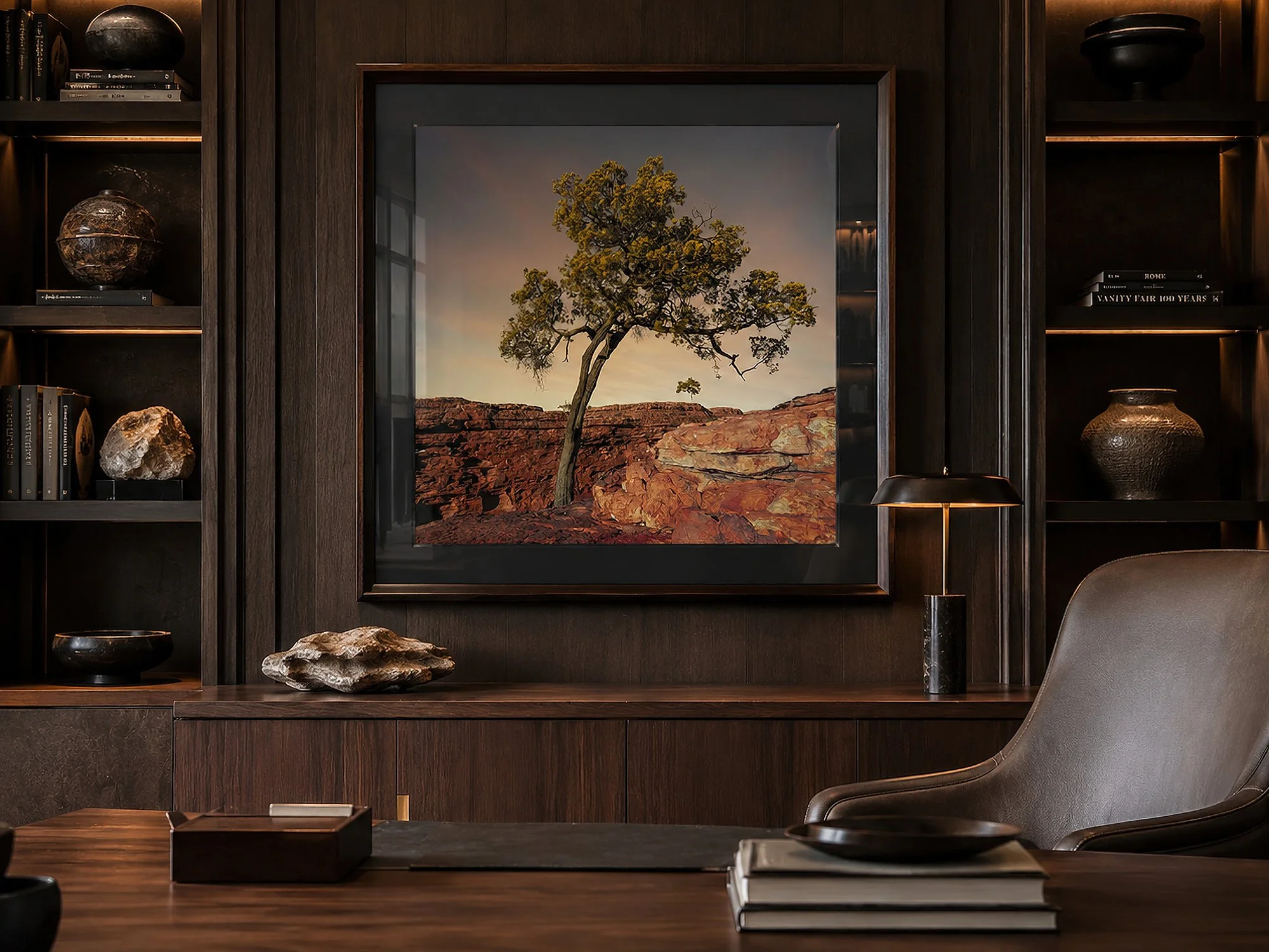 Dark wood-framed photograph of a solitary tree above red canyon rock, displayed with a dark mat in a moody luxury study with warm timber shelving, sculptural stone accents and soft ambient lighting, by Nick Psomiadis at NIKART Studio and Gallery