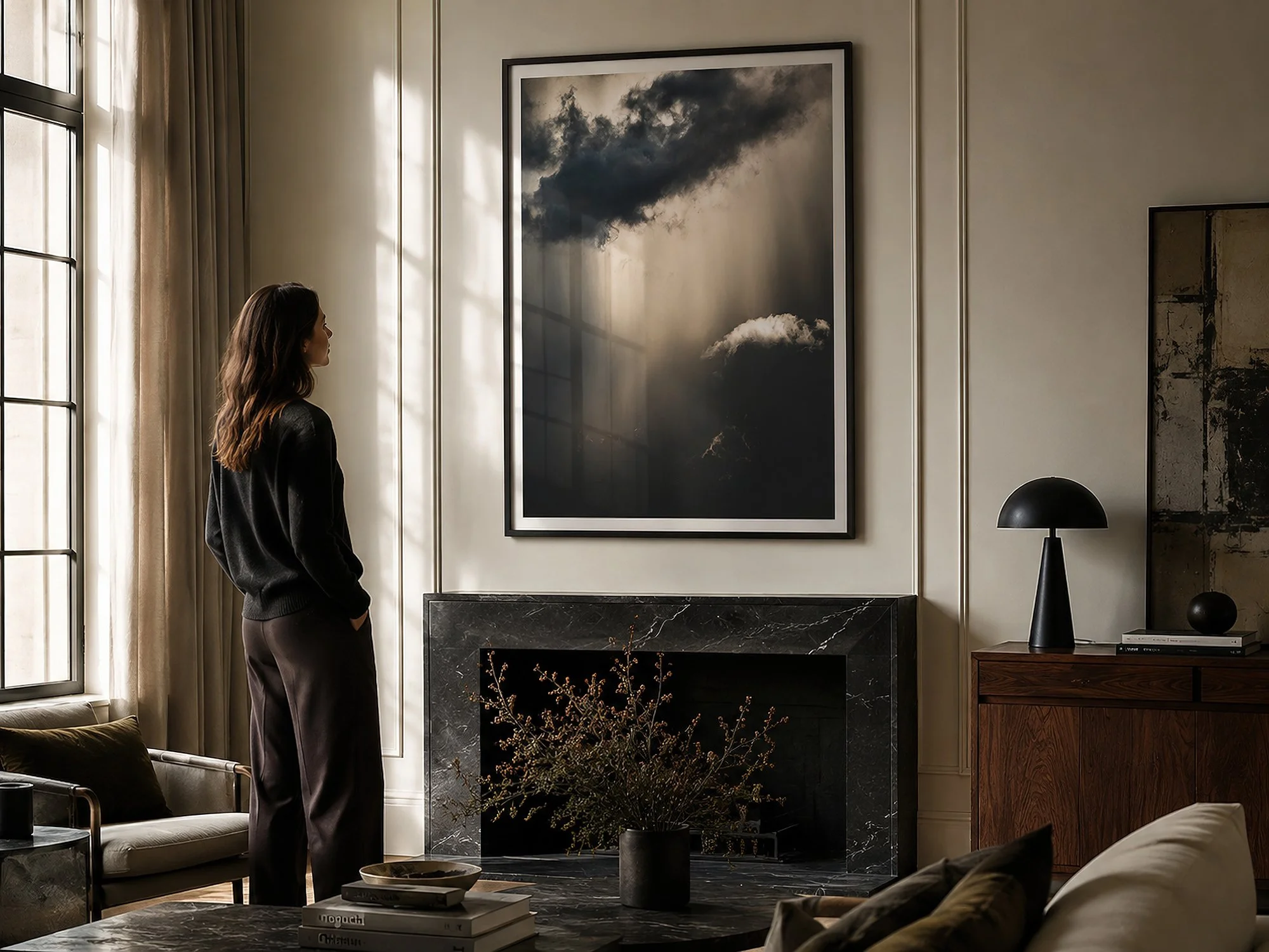 Bright moody living room with a woman viewing a black-framed dramatic cloud photograph above a marble fireplace, with natural light and glass reflections by Nick Psomiadis at NIKART Studio and Gallery