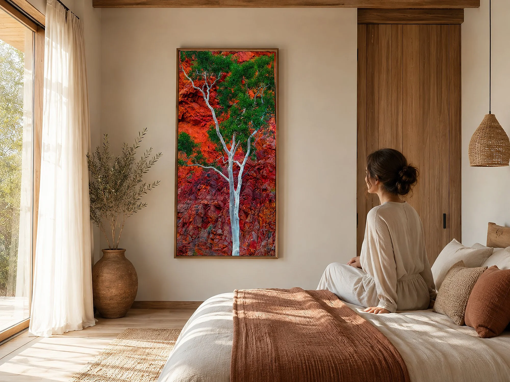 Wood-framed canvas of a white gum with green foliage against vivid red rock in a sunlit minimalist bedroom with warm timber, natural textures and a seated woman, by Nick Psomiadis at NIKART Studio and Gallery