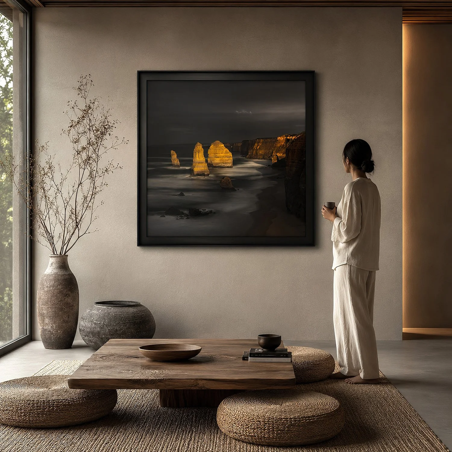 Square framed photograph of illuminated Twelve Apostles displayed in a warm minimalist interior with natural textures and a woman holding a cup, by Nick Psomiadis at NIKART Studio and Gallery