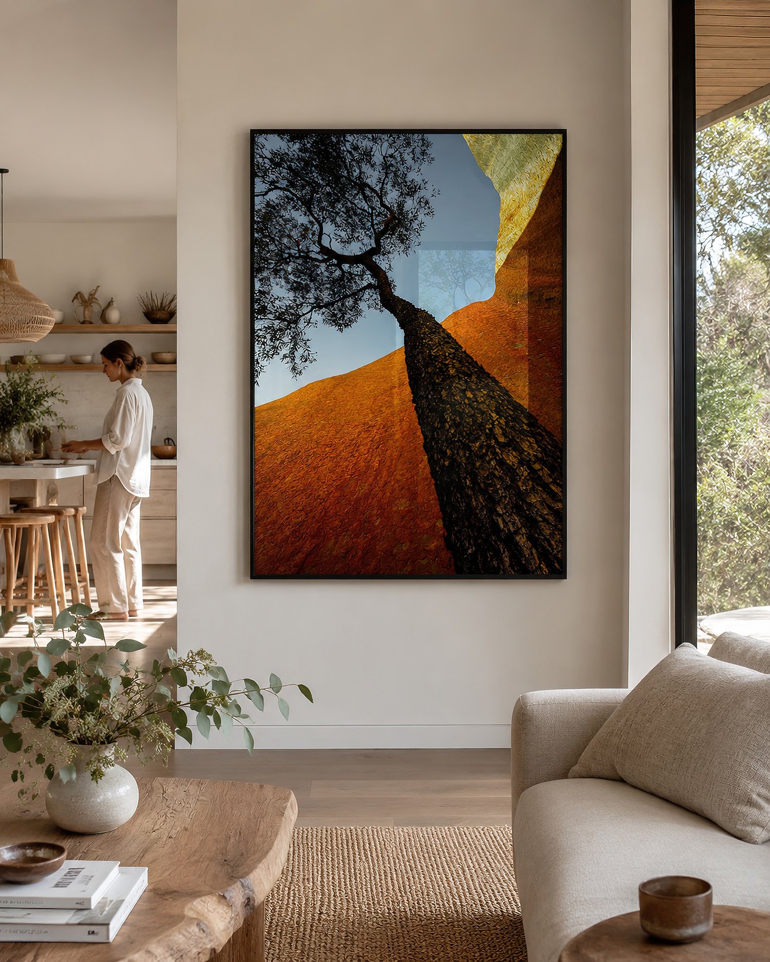Black-framed photograph of Uluru and a silhouetted tree with glass reflections in a naturally lit Australian living room, with a person standing in the adjoining kitchen, by Nick Psomiadis at NIKART Studio and Gallery