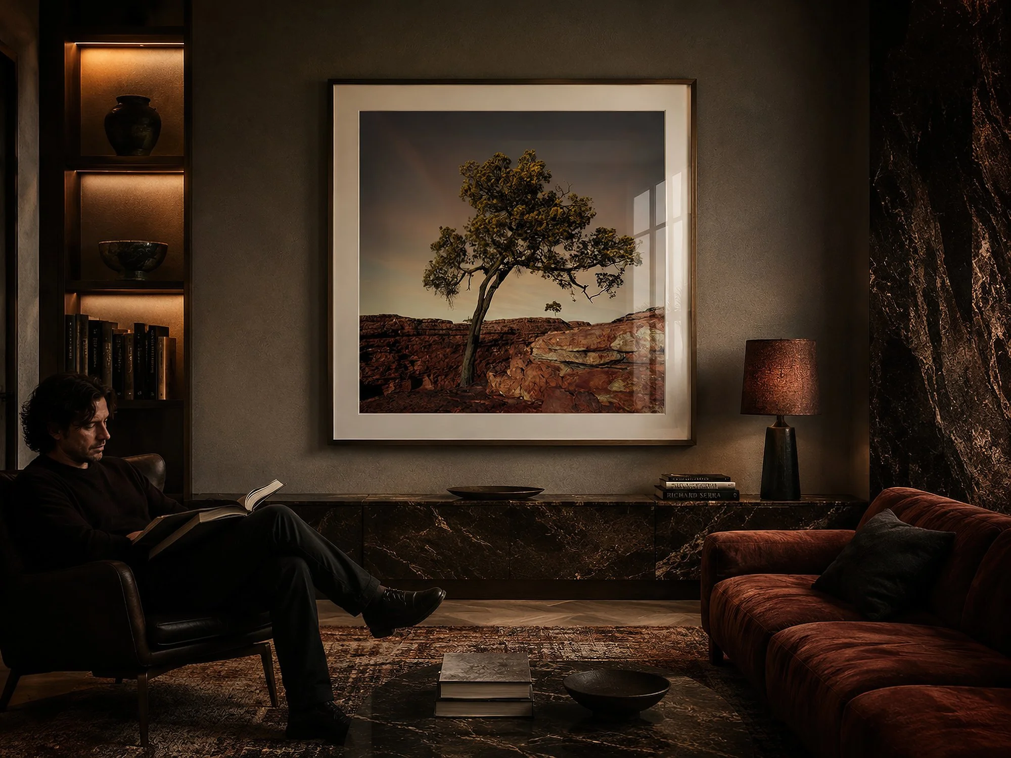 Framed Kings Canyon photograph of a solitary native tree above layered red sandstone in a moody luxury living room with dark timber, marble, warm lighting and a seated man reading, by Nick Psomiadis at NIKART Studio and Gallery
