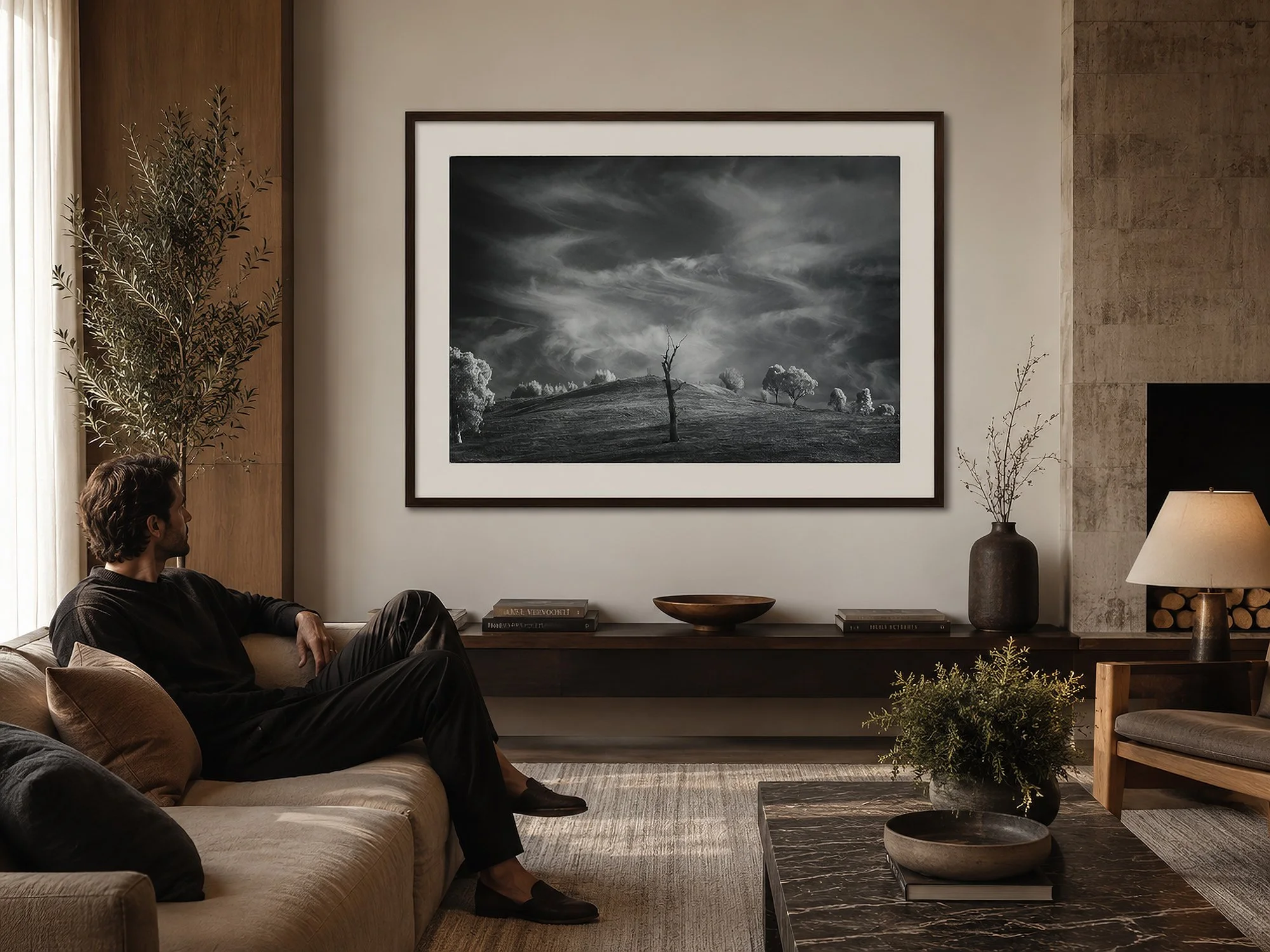 Large framed black-and-white landscape photograph with white mat and dark timber frame in a luxurious living room, with a seated man, neutral sofa, marble coffee table and warm contemporary décor by Nick Psomiadis at NIKART Studio and Gallery