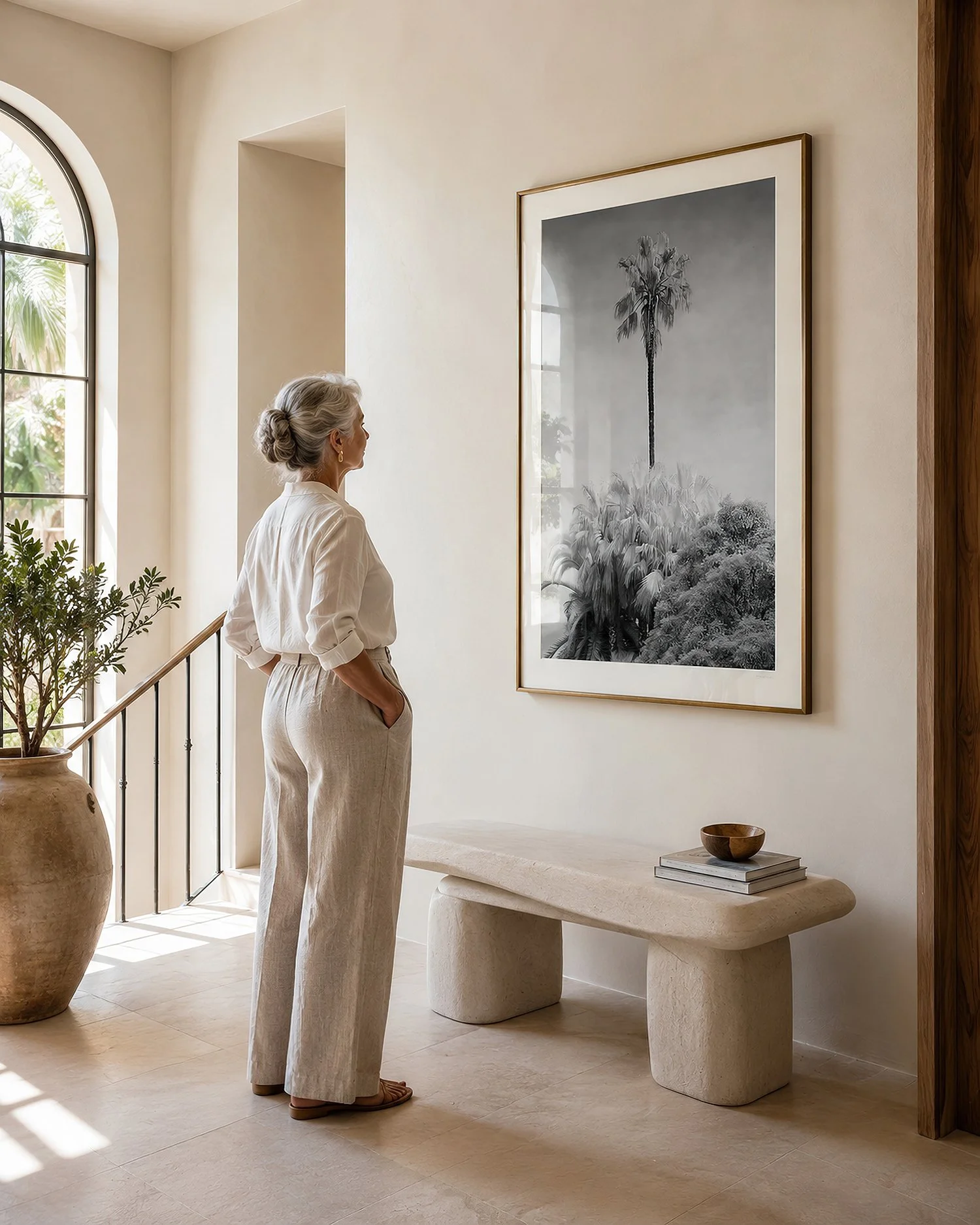 Bright minimalist hallway with an older woman viewing a gold-framed monochrome palm tree photograph with glass reflections by Nick Psomiadis at NIKART Studio and Gallery