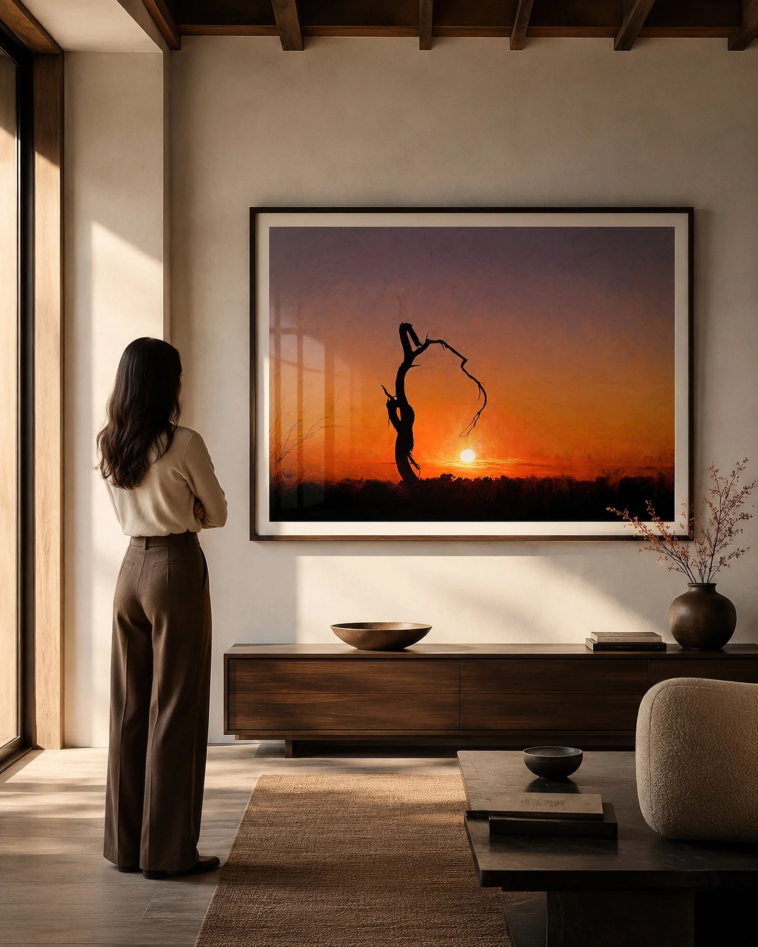 Bright moody living room with a woman viewing a large black-framed sunset silhouette photograph above a dark timber sideboard, with natural light and glass reflections by Nick Psomiadis at NIKART Studio and Gallery