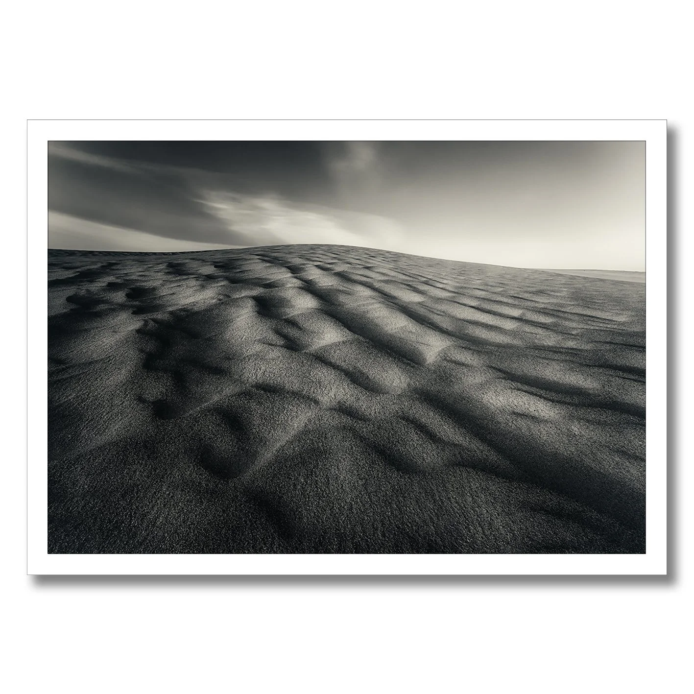 Black and white photograph of wind-sculpted sand dunes beneath a dramatic sky.