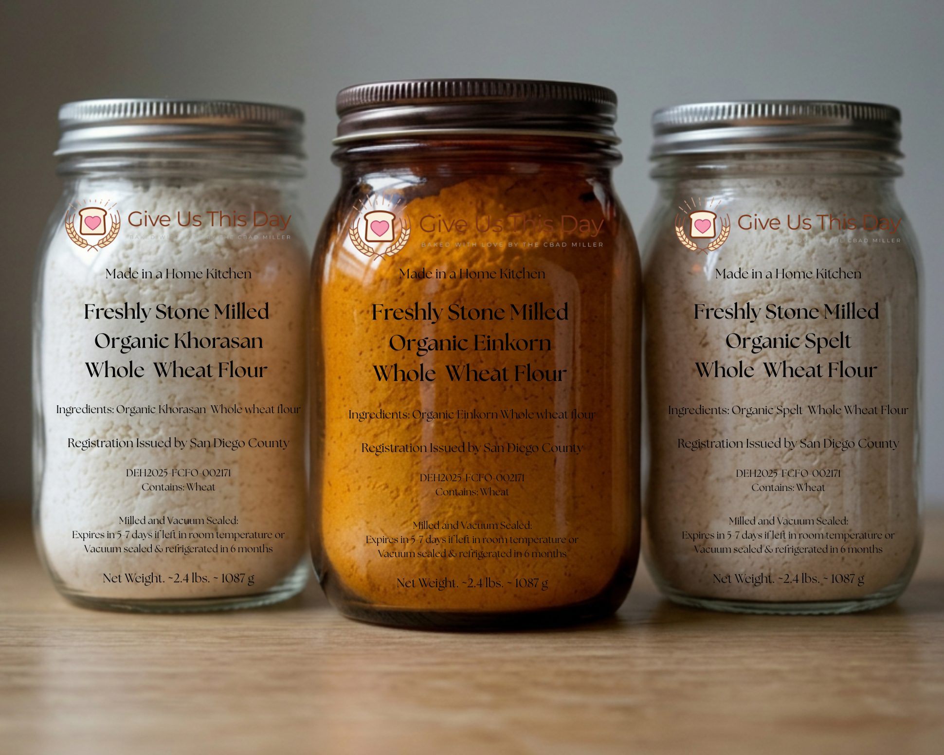 Three glass jars containing different types of flour labeled in black text. The left jar contains white flour labeled as "Freshly Stone Milled Organic Khorasan Whole Wheat Flour," the middle jar contains yellow-orange flour labeled as "Freshly Stone 