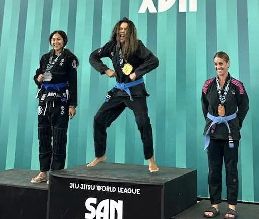 Three female Blue belt Brazilian Jiu-Jitsu competitors in a WJJL tournament on a podium enjoying the moment. The winner exuberates joy mid air.